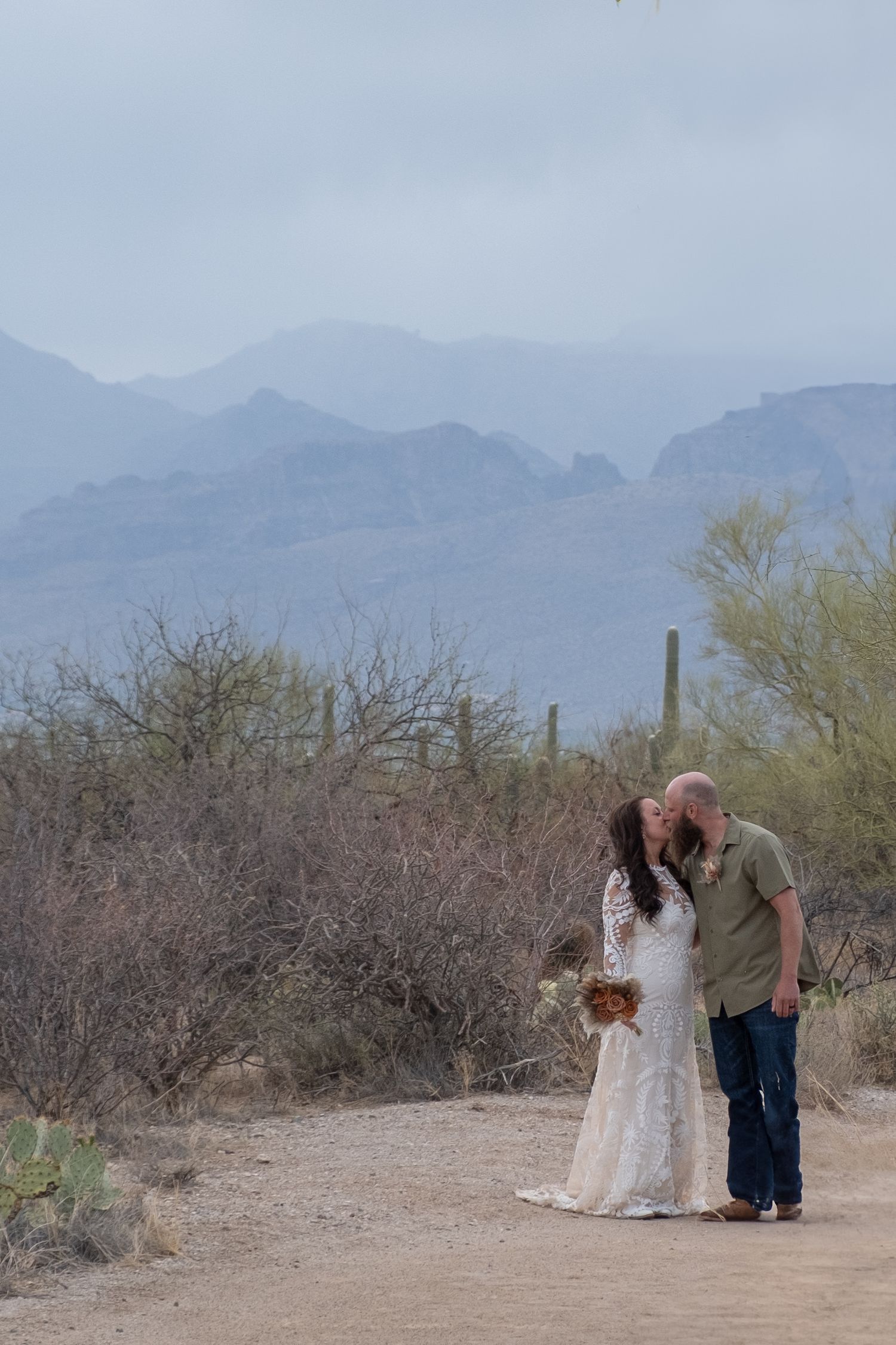 Two people share an intimate moment on a desert trail with saguaro cacti and misty mountains in the background during their elopement.