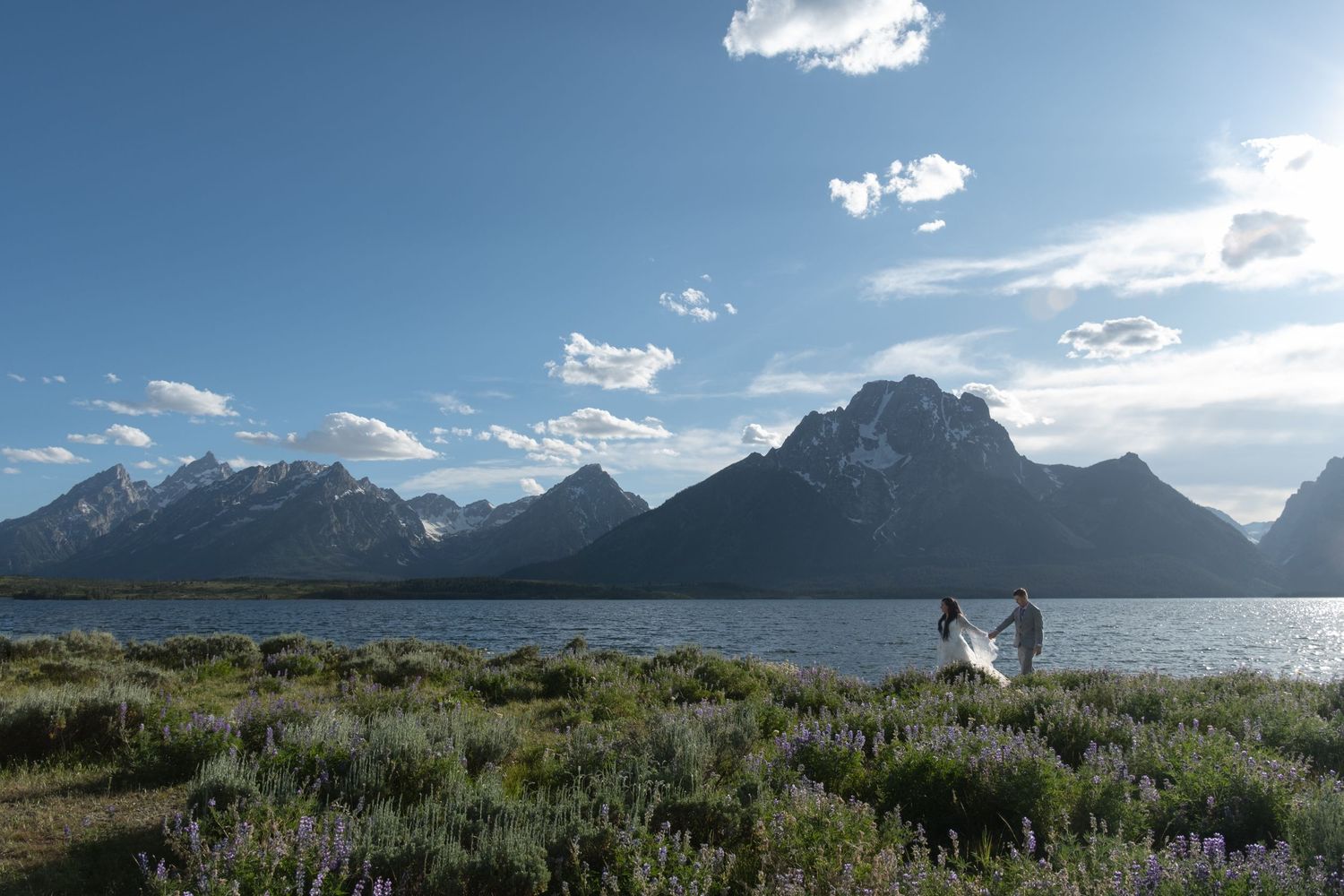 Two people stand on grassy shoreline overlooking Grand Teton mountains and Jackson Lake under blue skies in Wyoming.