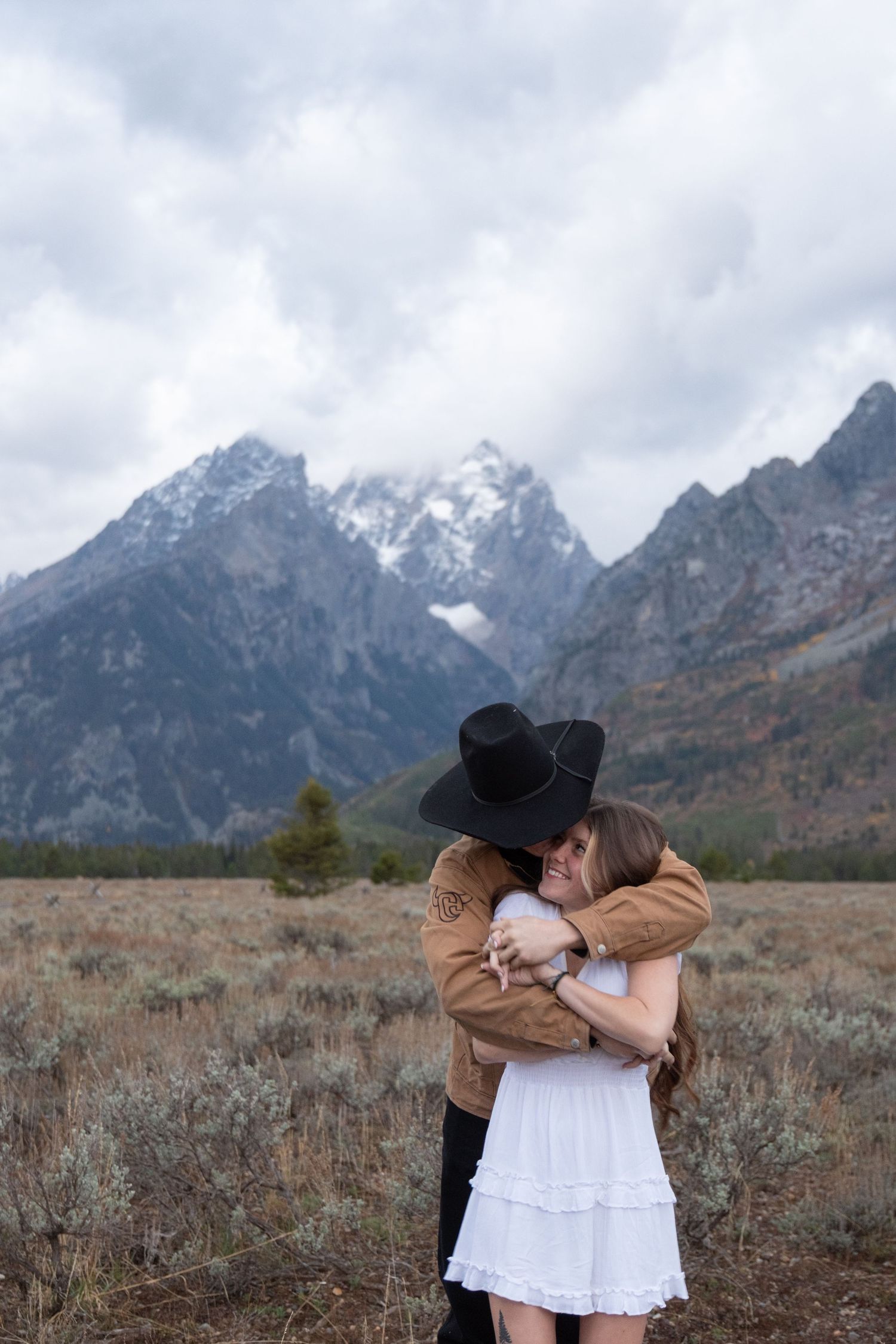 A couple embraces in front of snow-capped mountains at Grand Teton National Park on an overcast day.