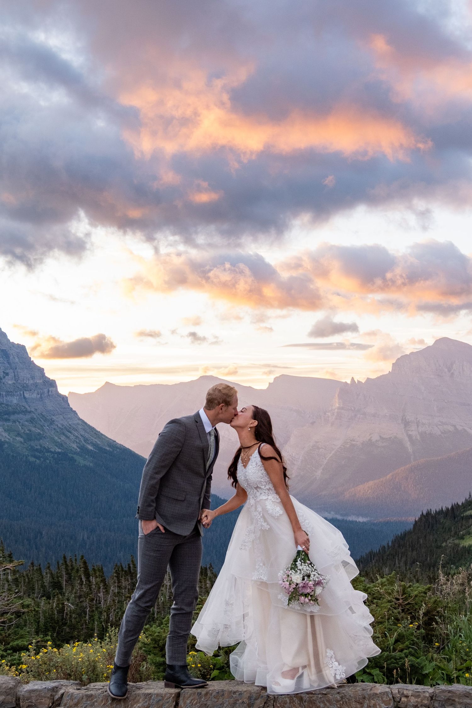 A couple shares a romantic kiss against a backdrop of majestic mountain peaks and dramatic sunset clouds.