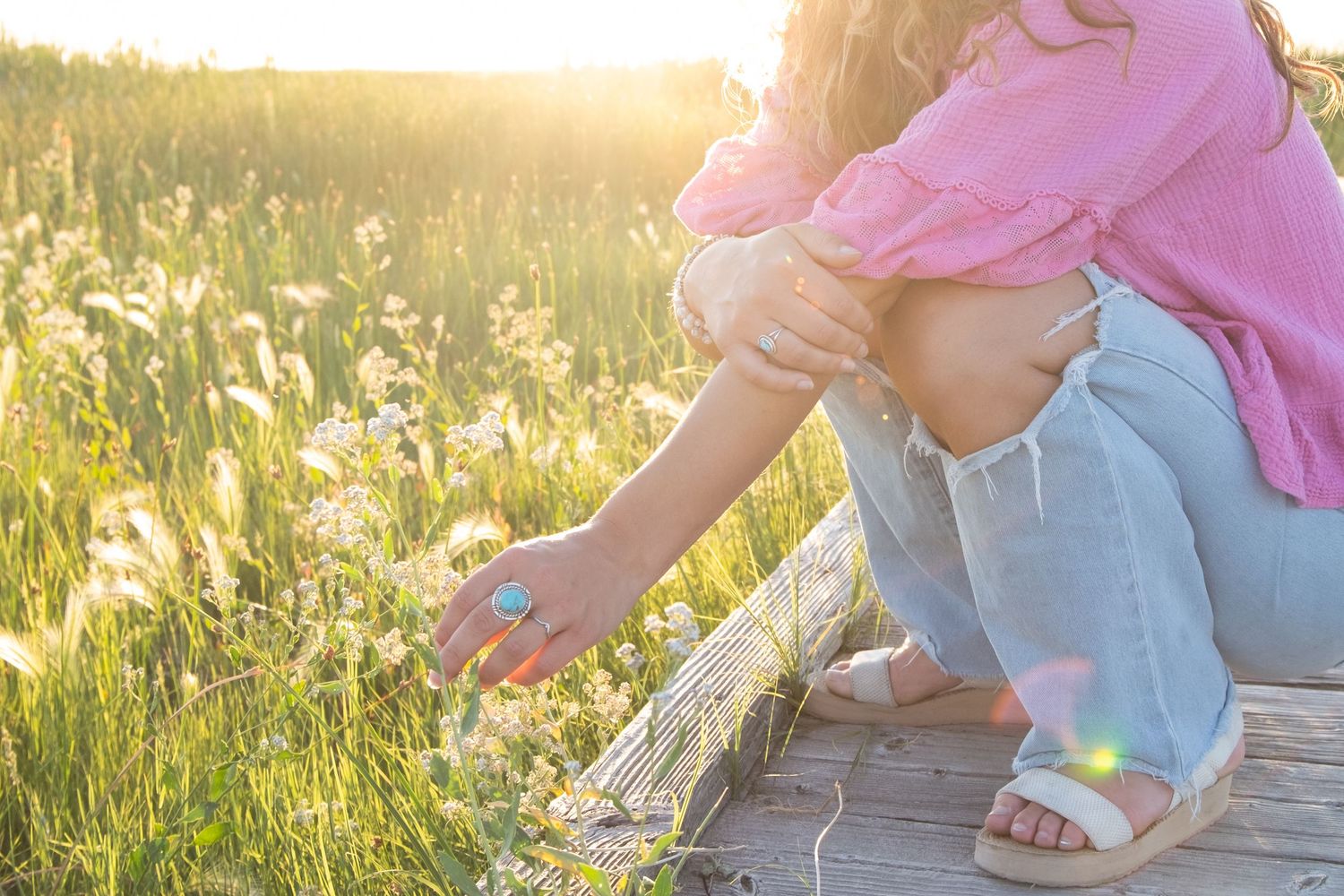 A person in jeans and pink shirt crouches in a wildflower meadow at golden hour reaching for flowers.