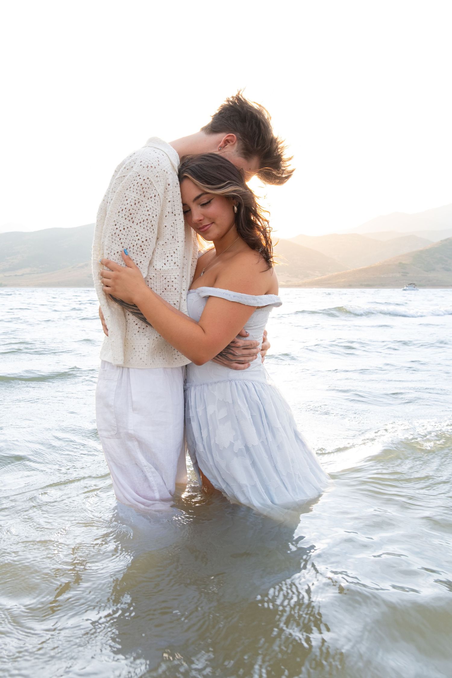 A couple embraces in ocean waves at sunset while wearing white and light blue romantic beach attire.