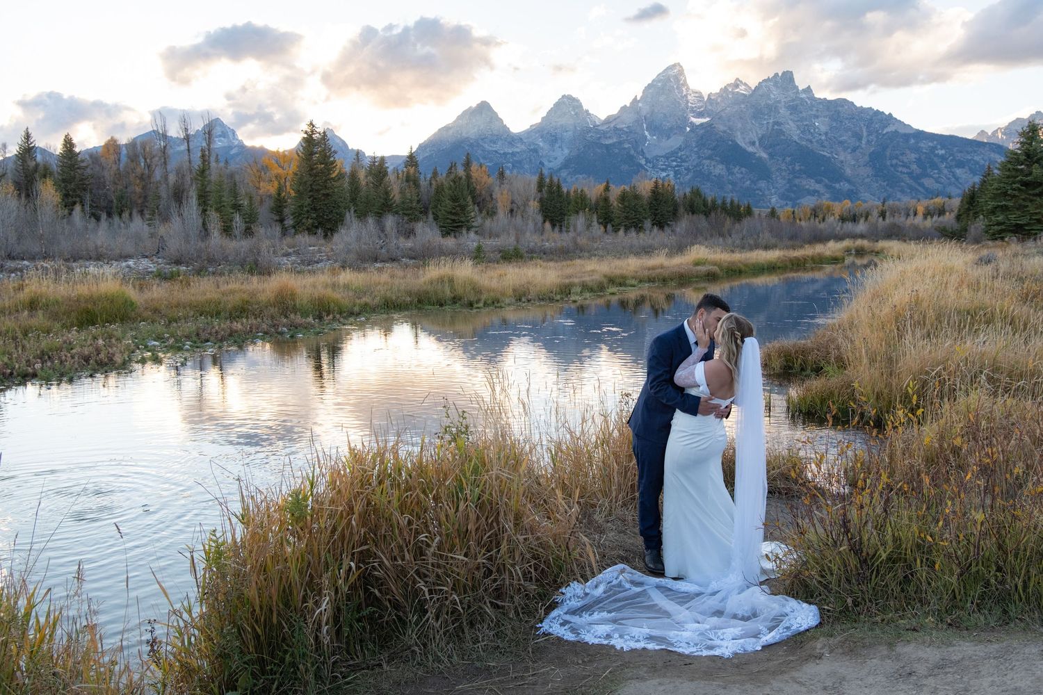 A couple in wedding attire embraces near a scenic mountain lake at sunset with dramatic peaks in the background.