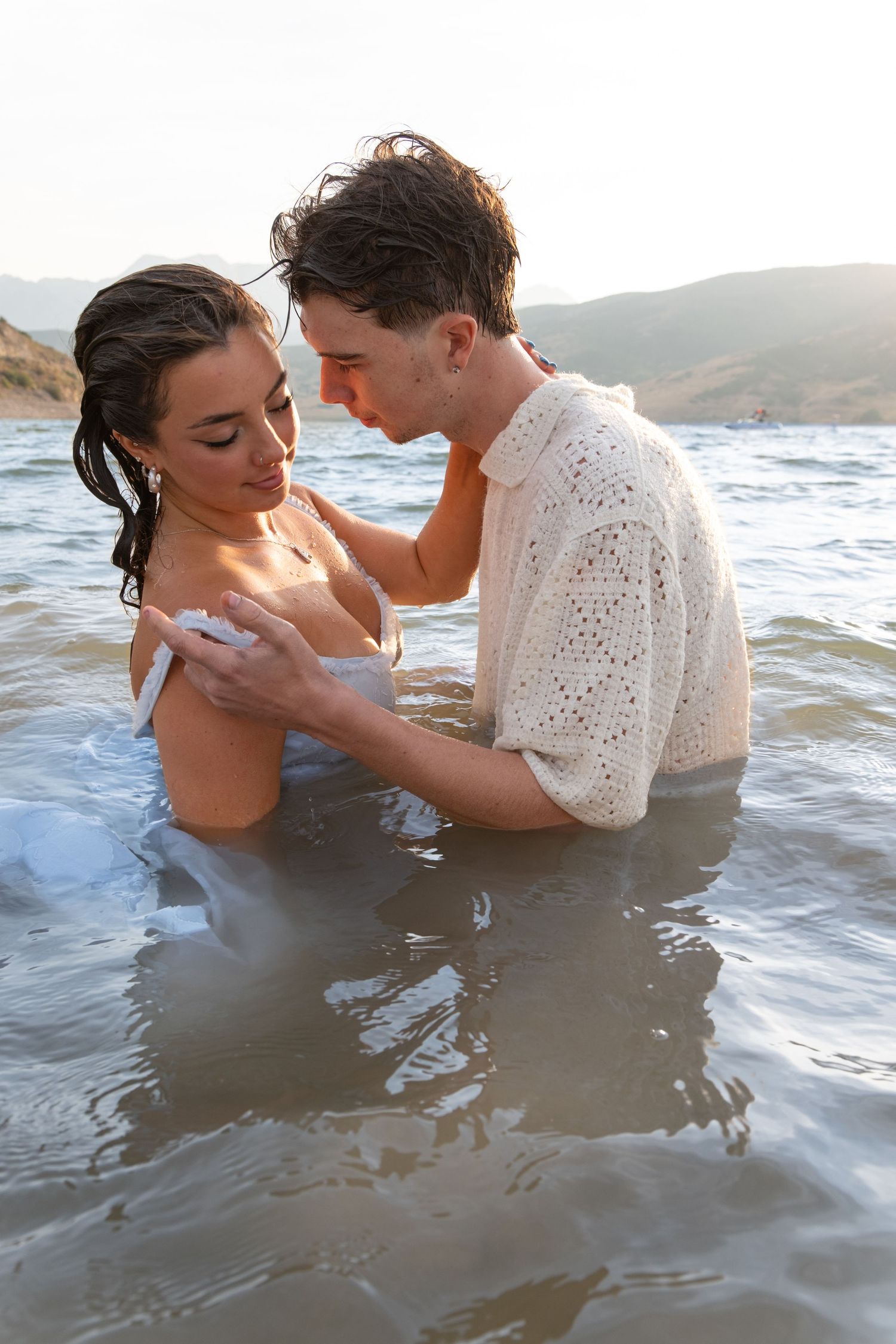 A romantic couple embraces while wading in ocean water on a sunny day with hills in the background.