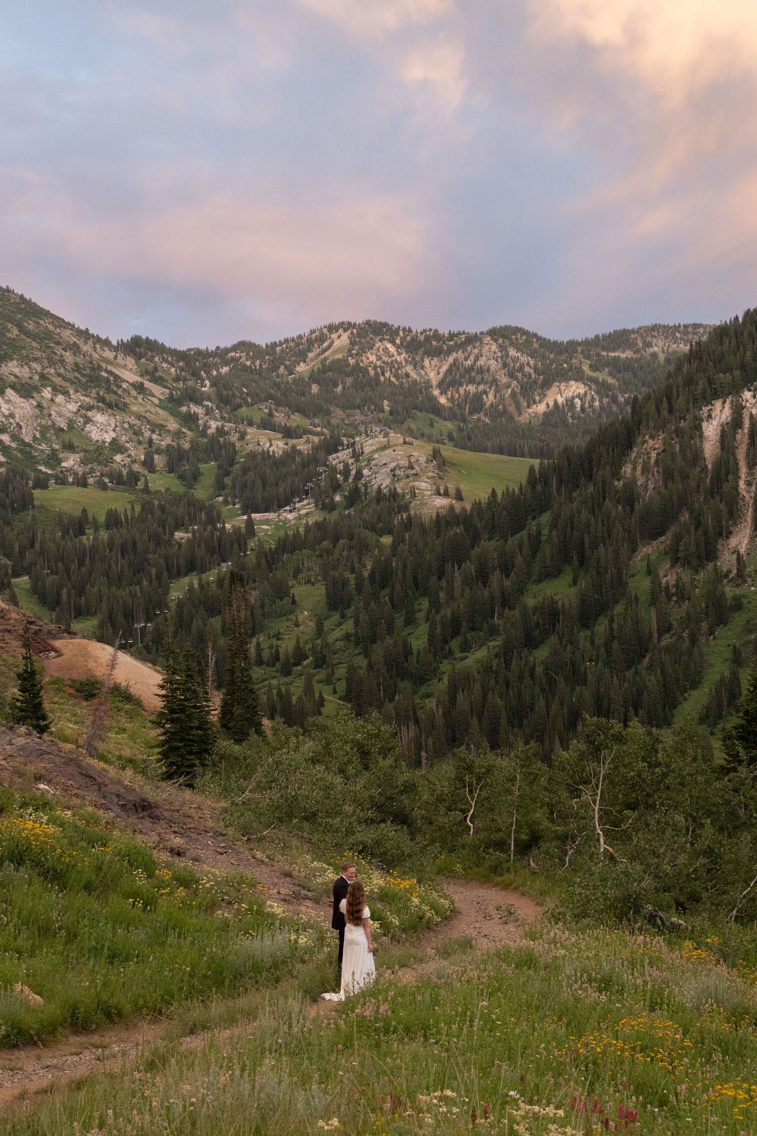 Two people walk through a scenic mountain meadow with wildflowers and evergreen trees against a sunset sky.