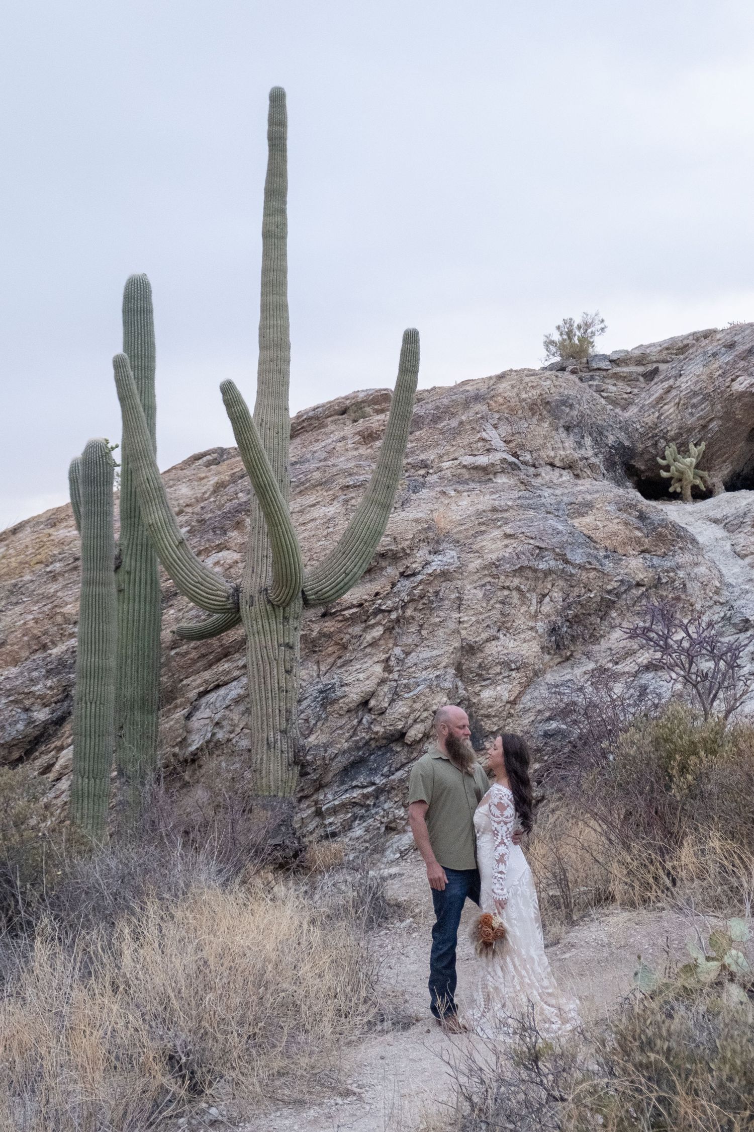 A couple embraces in front of towering saguaro cacti and rocky desert landscape during their elopement photoshoot.