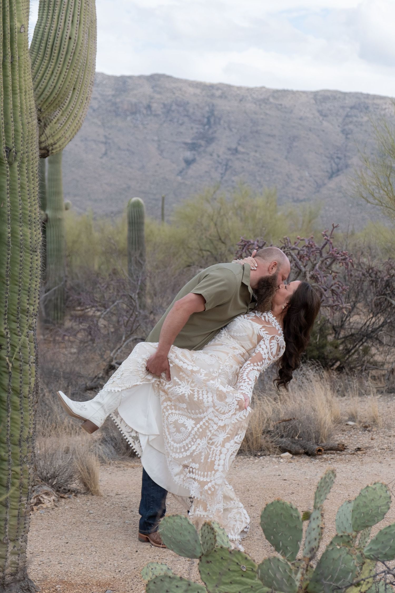A romantic couple embraces in front of towering saguaro cacti and mountains in the Arizona desert landscape during their elopement.