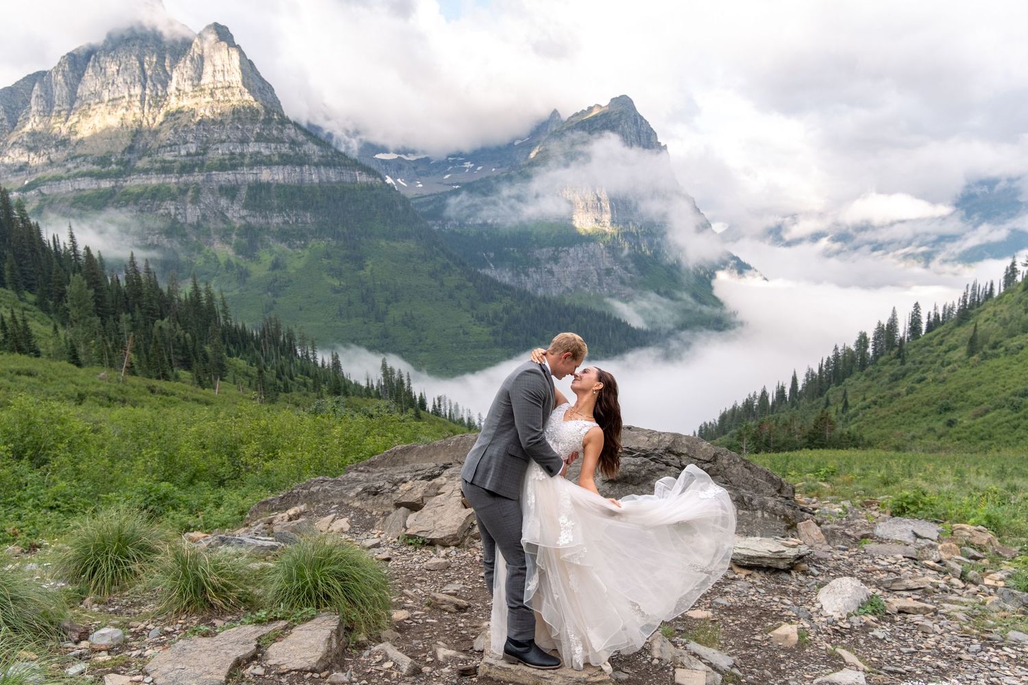 A couple in wedding attire embrace on a mountain overlook with misty peaks and green valleys in Glacier National Park.