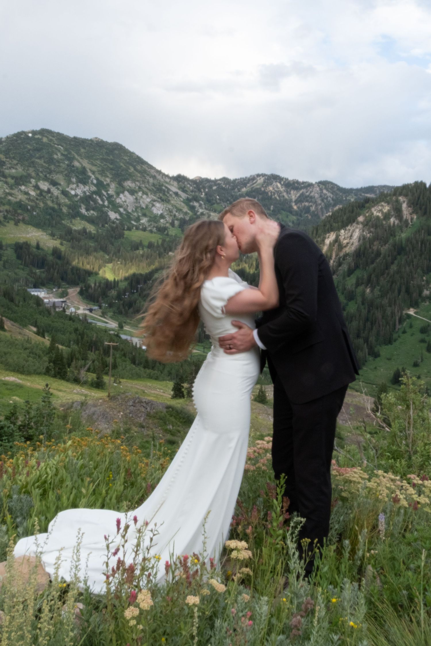 Newlyweds embrace against backdrop of rugged mountain landscape and summer wildflowers.