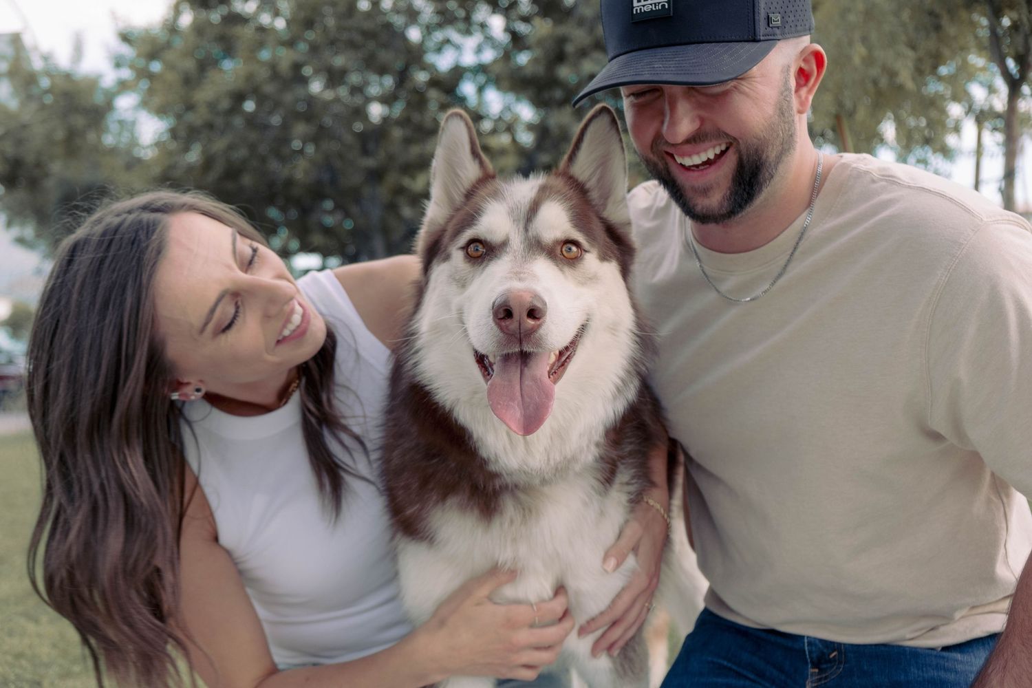 Couple playing with their smiling Husky dog in a grassy park setting.