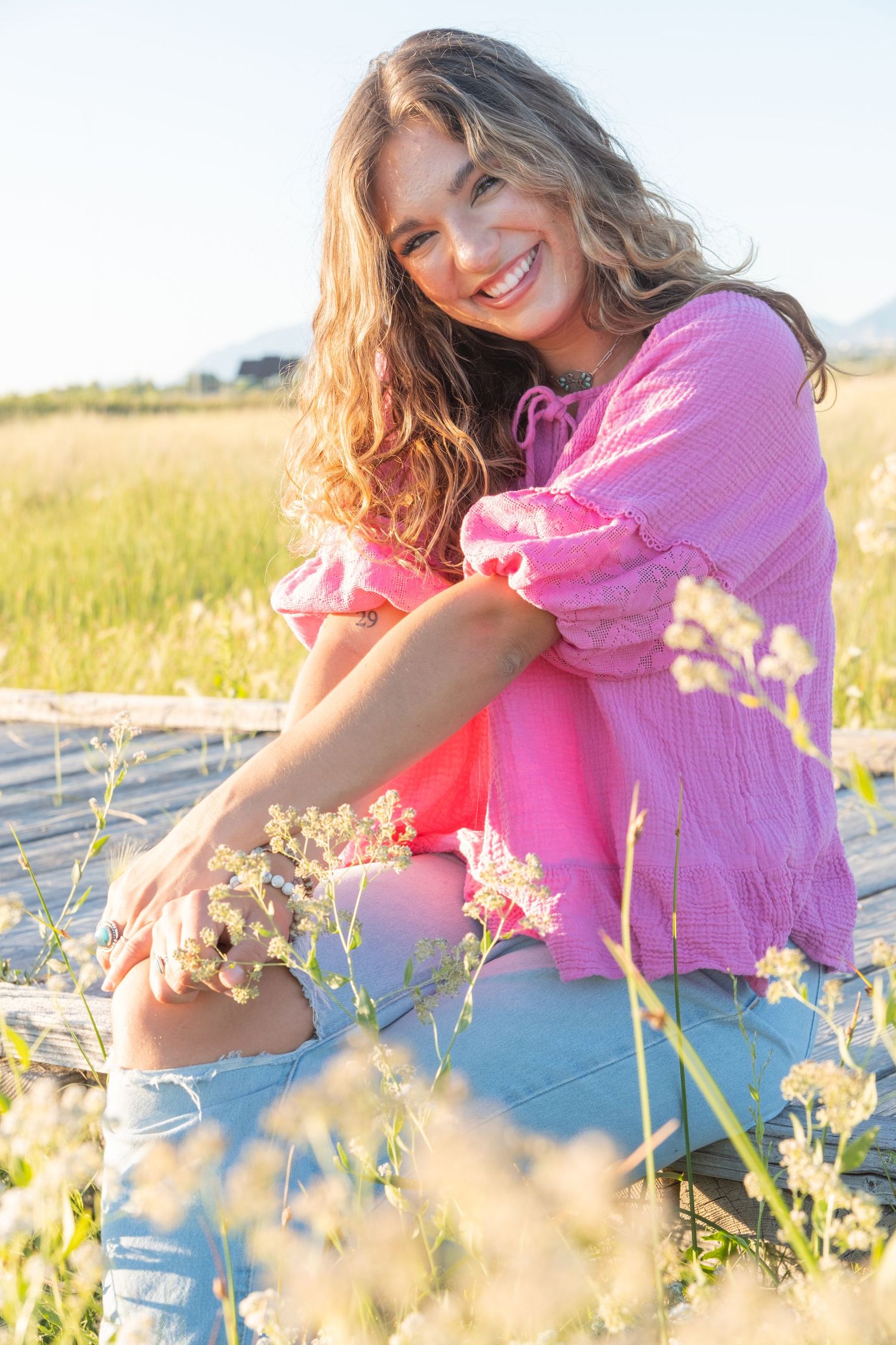 A person in a pink sweater sits among wildflowers in a sunny meadow during golden hour.