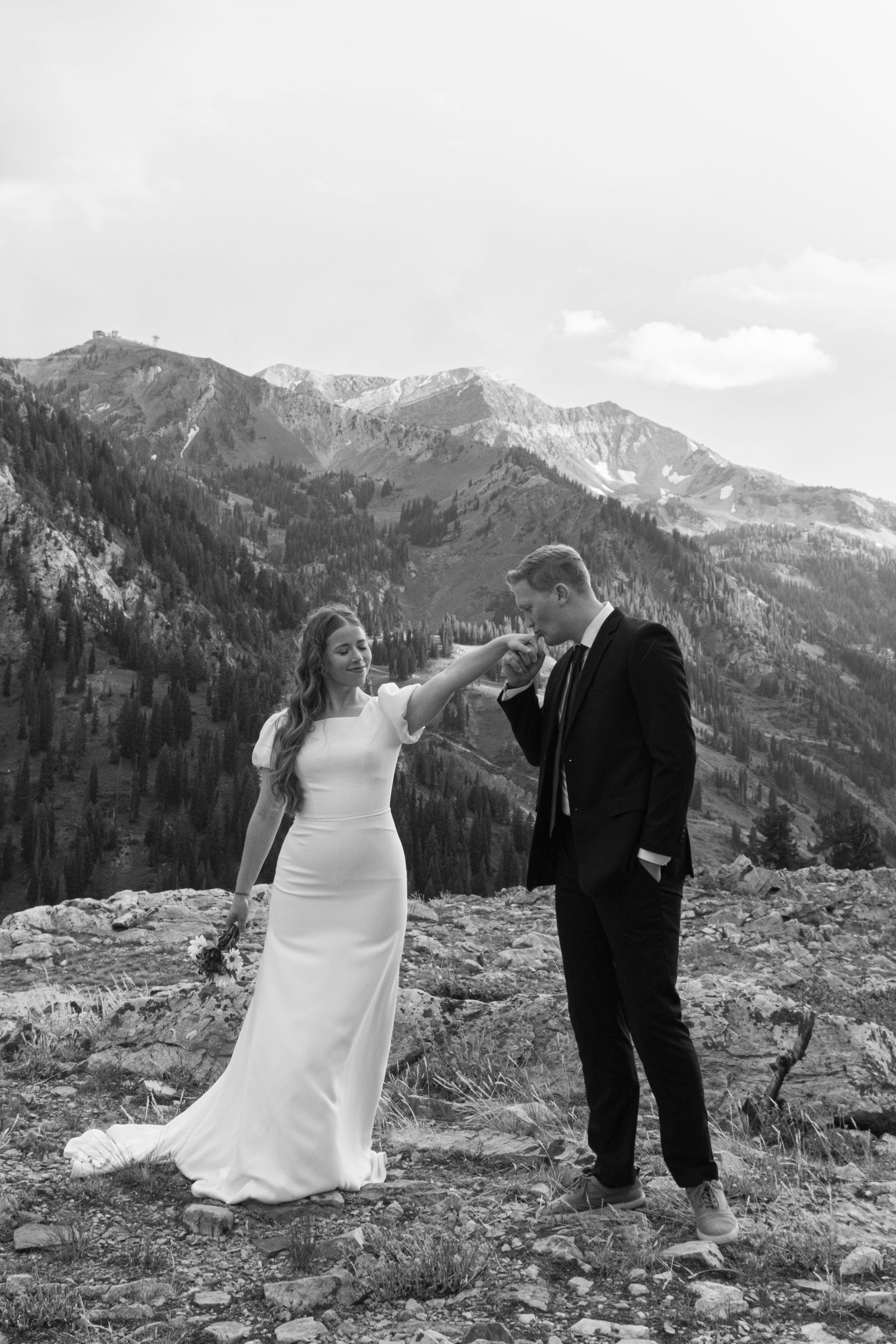 A couple in wedding attire embraces against a backdrop of snow-capped mountains in a black and white photograph.