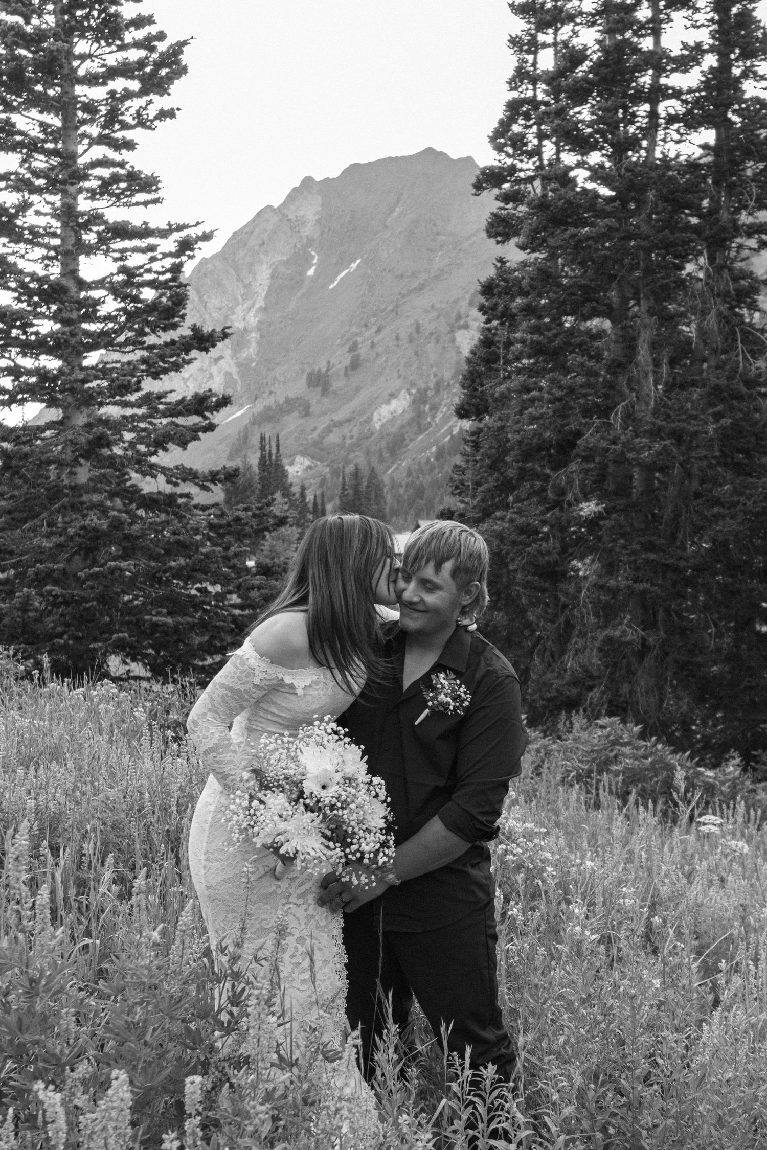 A couple embraces in a meadow with evergreen trees and a majestic mountain peak in the background.
