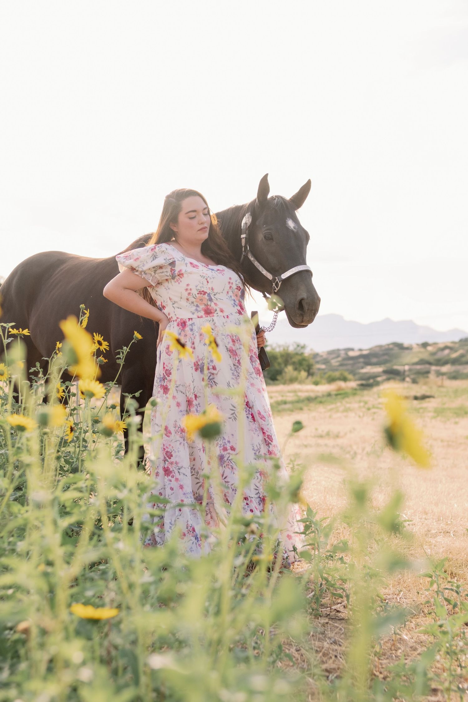 Person in a floral dress stands with a dark horse in a desert landscape with yellow wildflowers in the foreground.