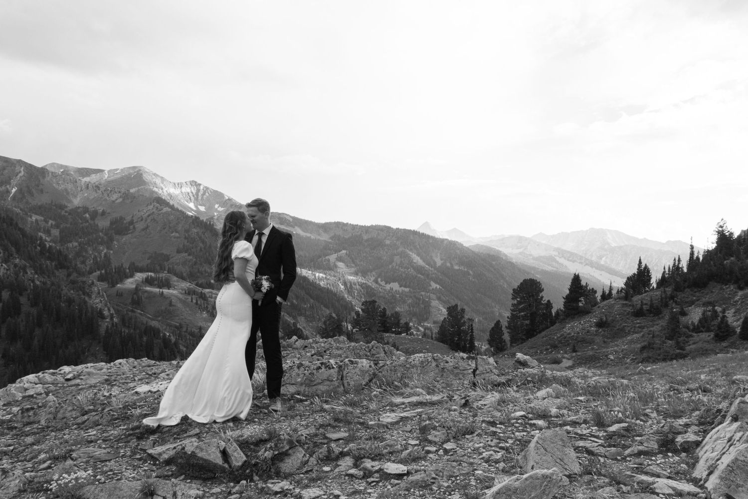 A couple stands together overlooking majestic snow-capped mountains in a scenic wilderness landscape.