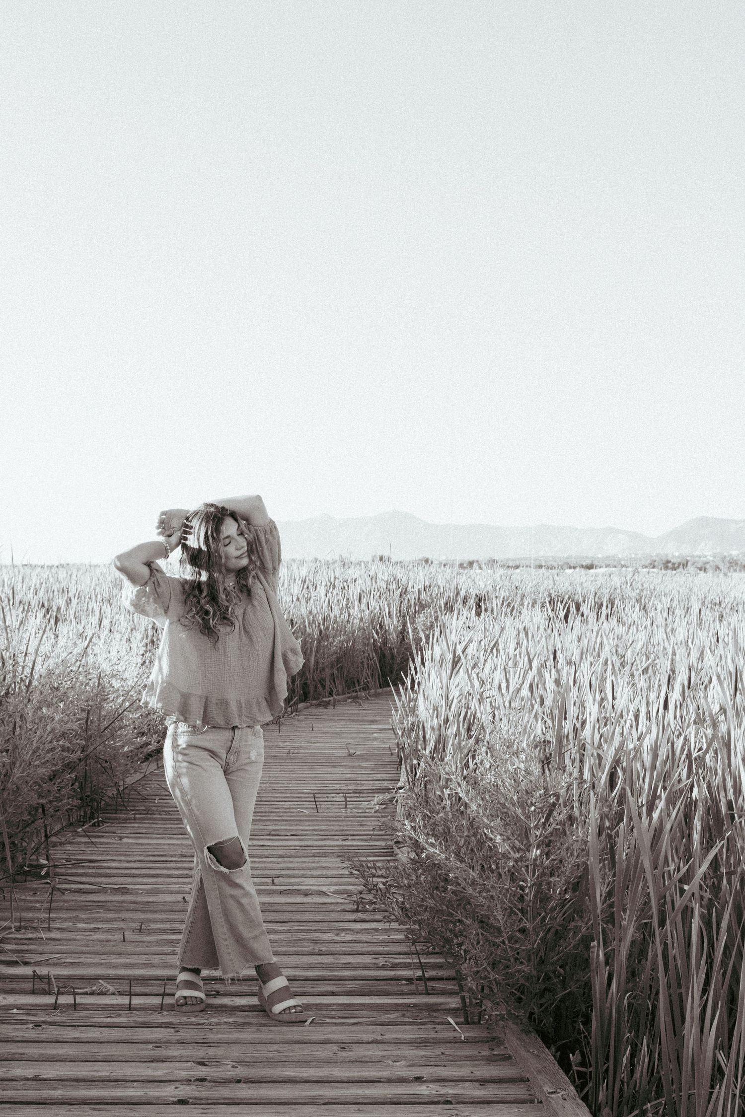 Figure in casual attire stands on wooden boardwalk path through tall reeds in monochrome landscape.