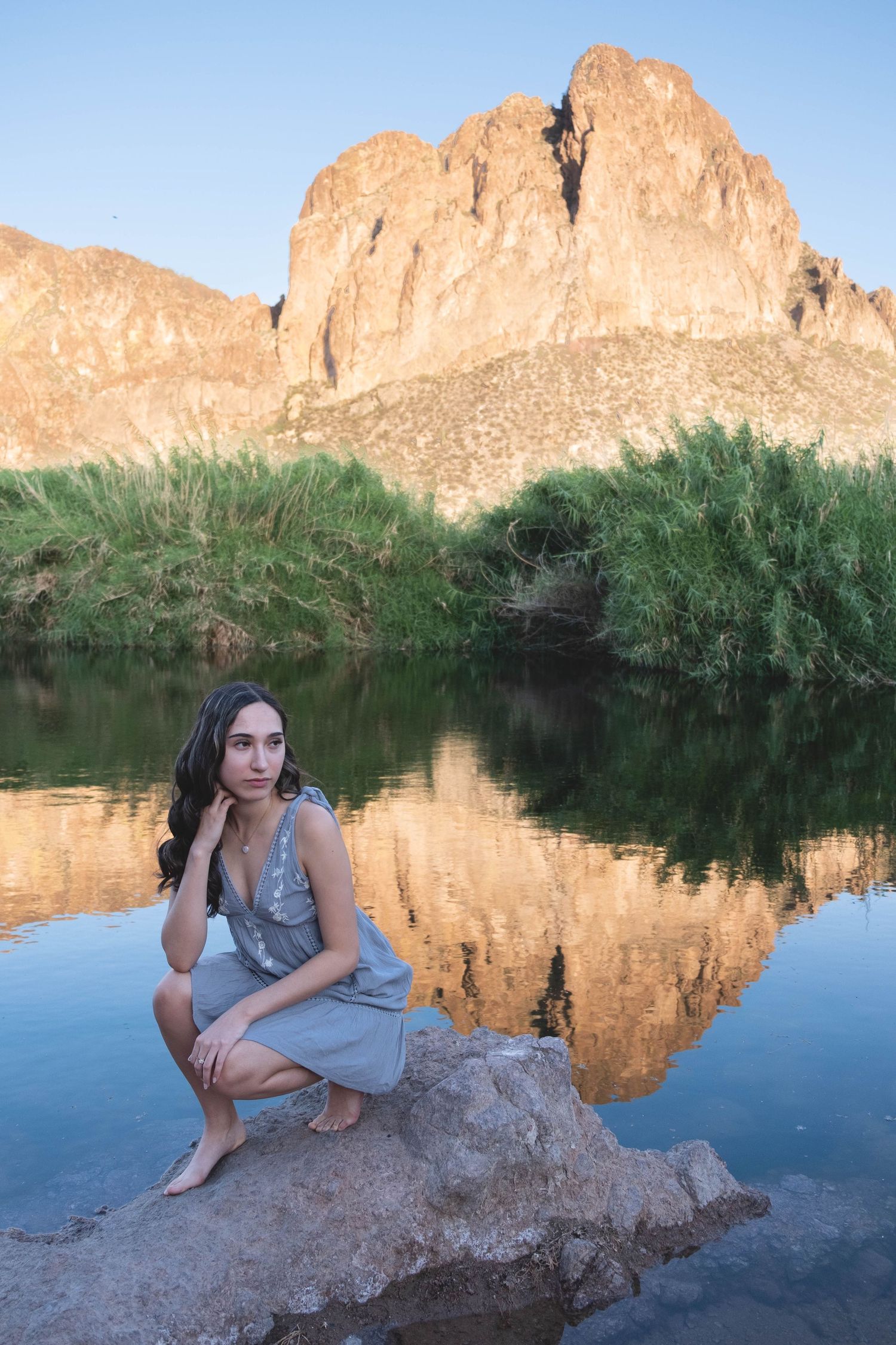 Desert mountains reflect in tranquil water as someone sits on a rock by the shoreline.