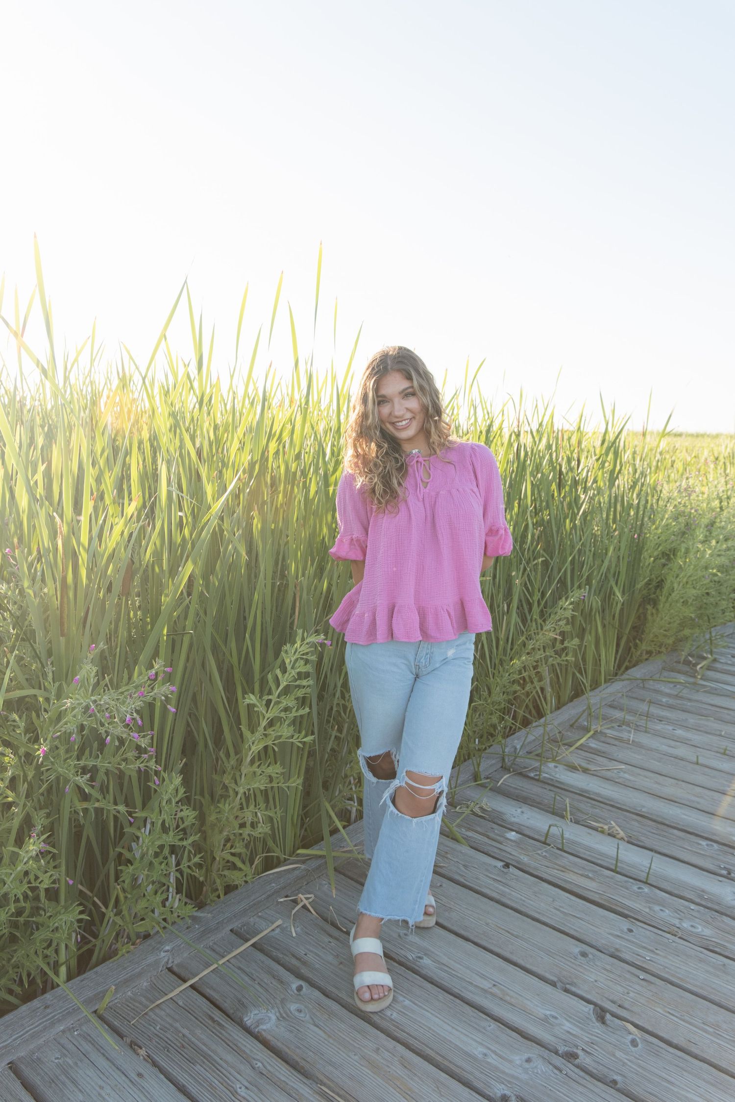 Person in pink blouse and ripped jeans poses on wooden boardwalk surrounded by tall grass at sunset.