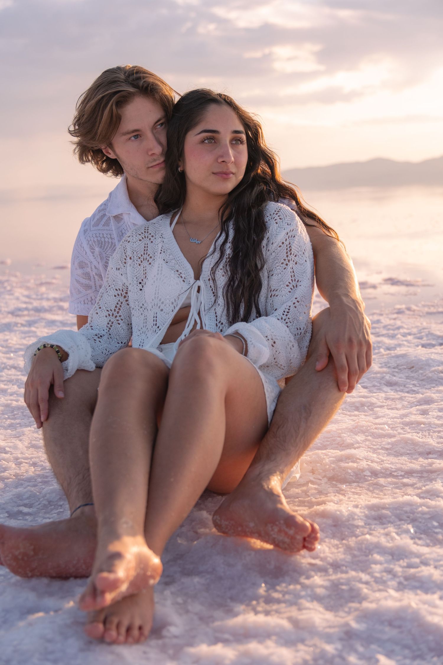 A couple in white lace outfits sits together on a salt flat at sunset, creating a romantic beachside atmosphere.