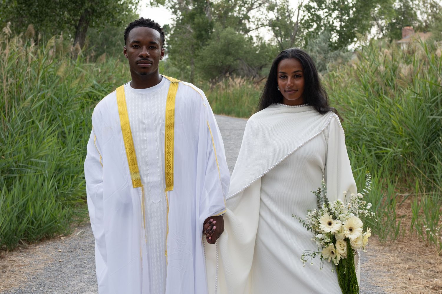 Couple in traditional white Ethiopian wedding attire standing on a path surrounded by lush greenery, holding a bouquet.