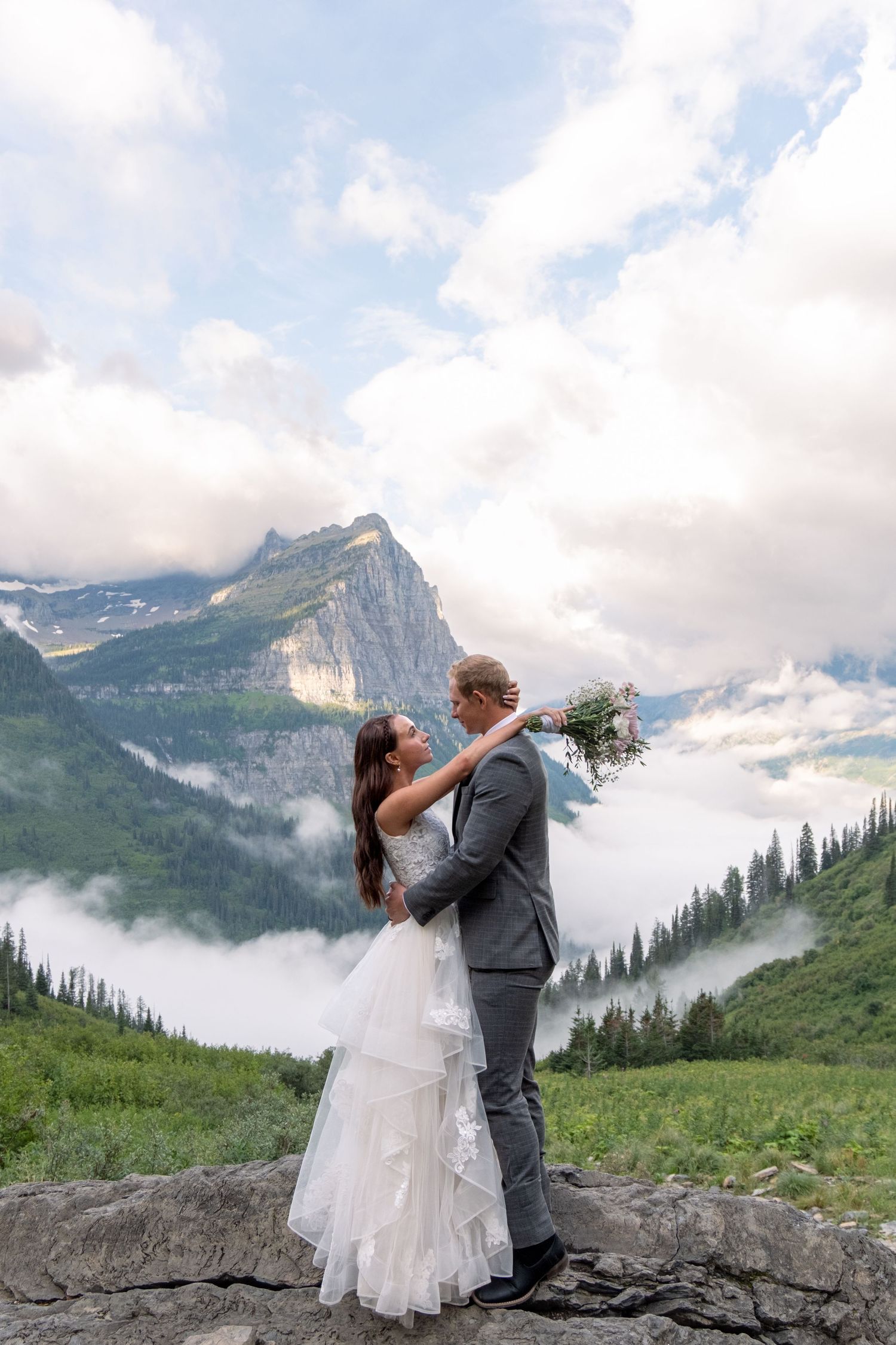 A couple in wedding attire embraces on a mountain overlook with dramatic peaks and misty valleys in Glacier National Park.