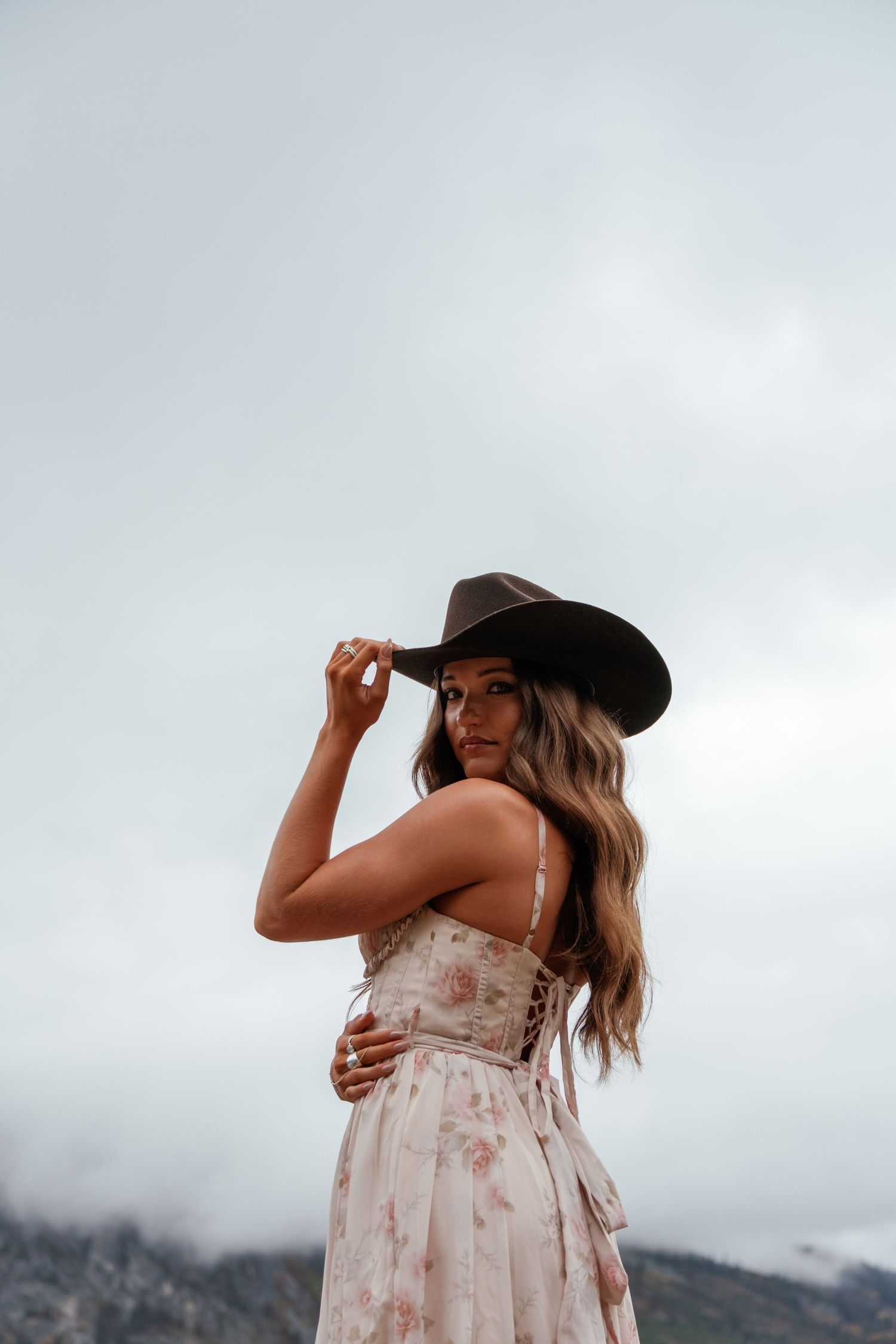A person in a floral dress and cowboy hat poses against a cloudy mountain landscape.