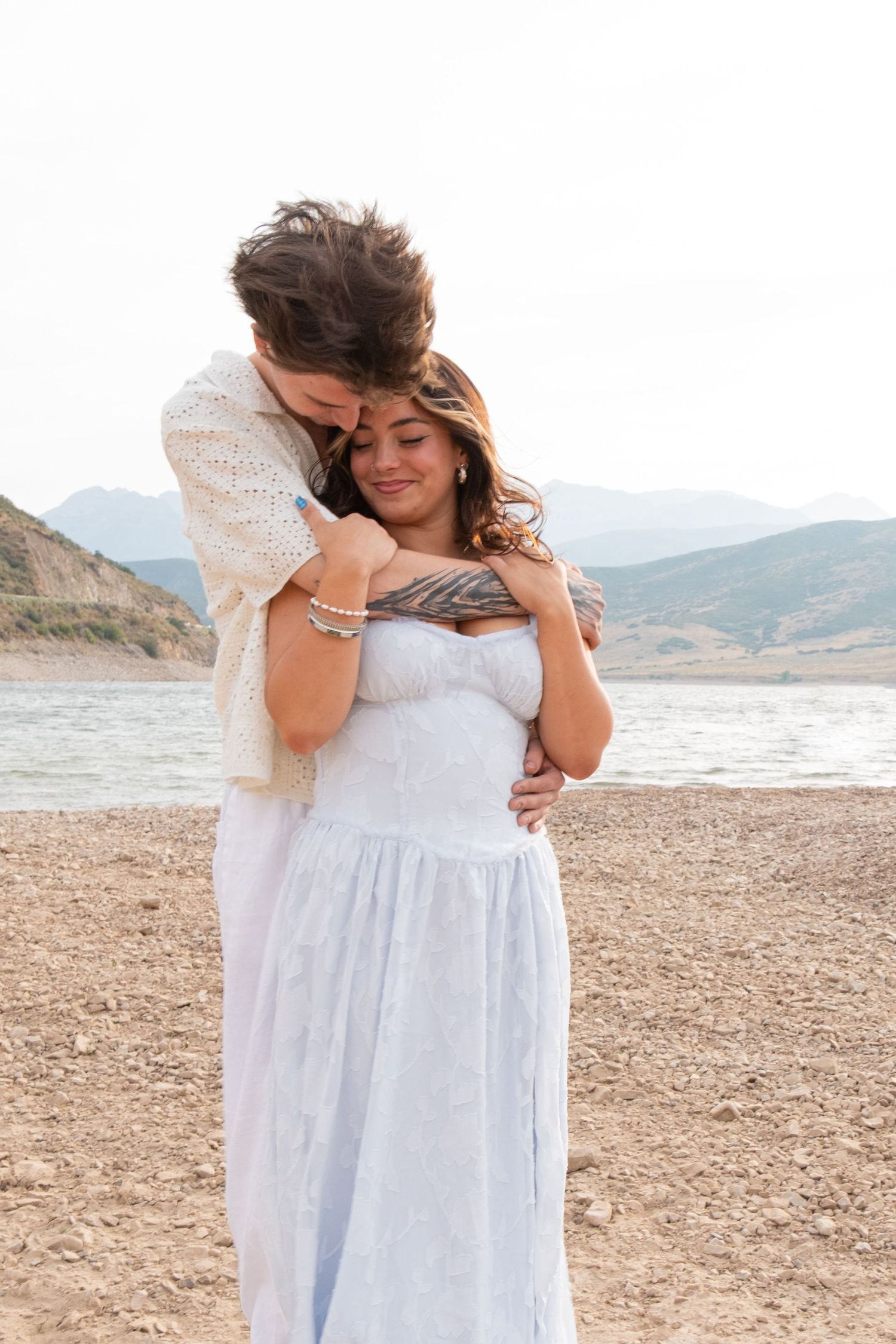 A person wearing a flowing white dress poses gracefully on a rocky beach with mountains in the background.