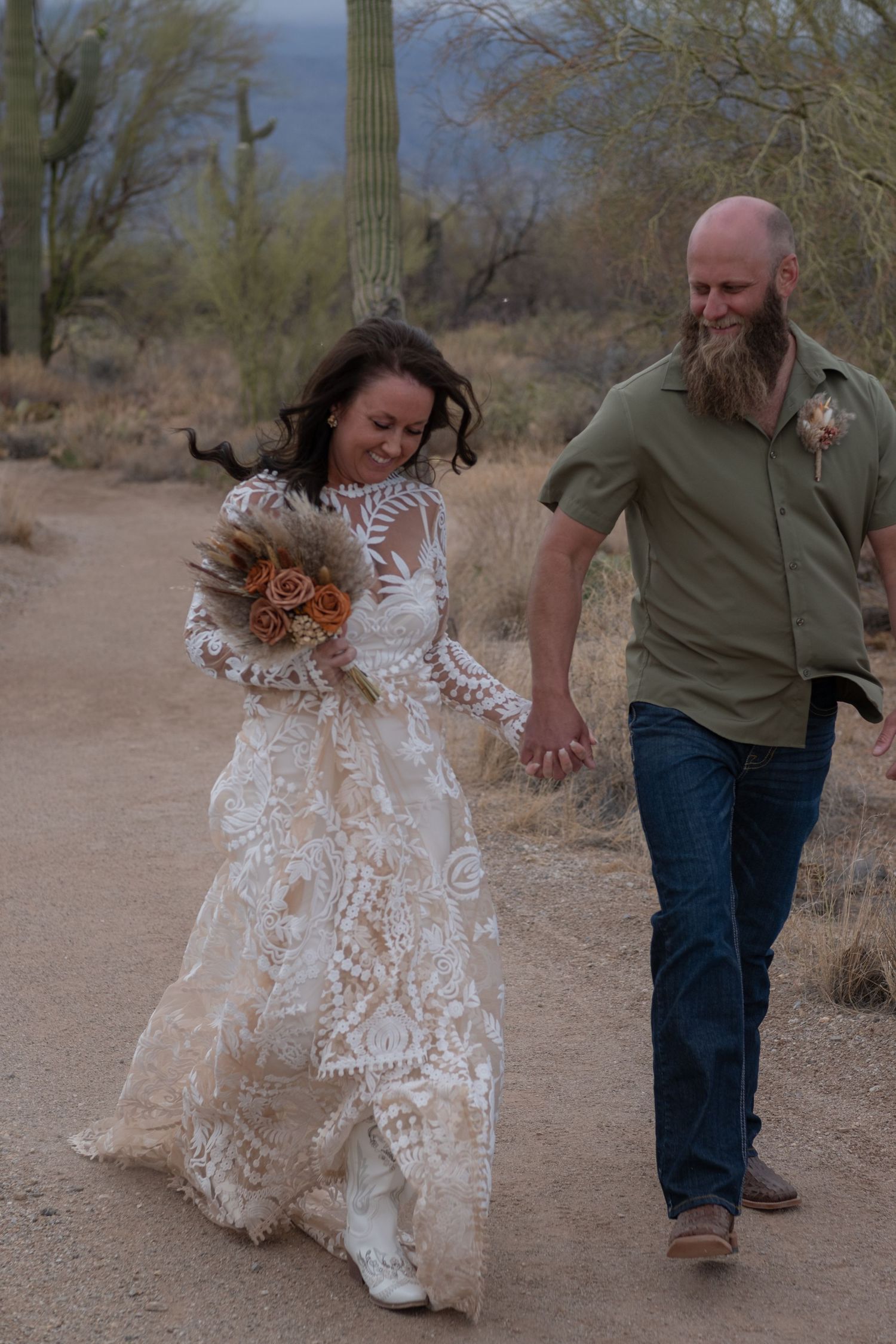 An eloping couple joyfully walking on a desert trail surrounded by saguaro cacti, with the bride wearing a lace wedding dress.