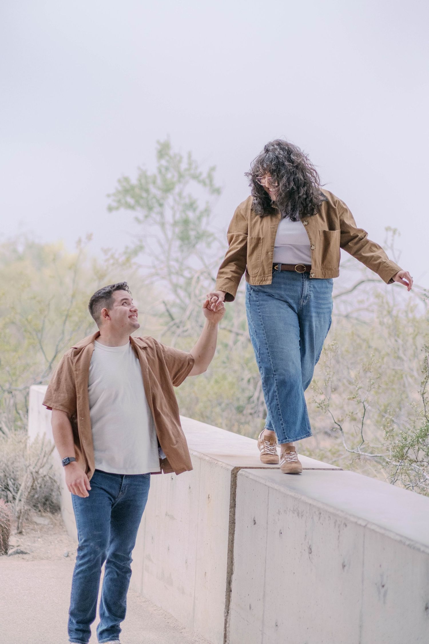 A couple playfully interacts on a concrete wall with desert scenery behind them.
