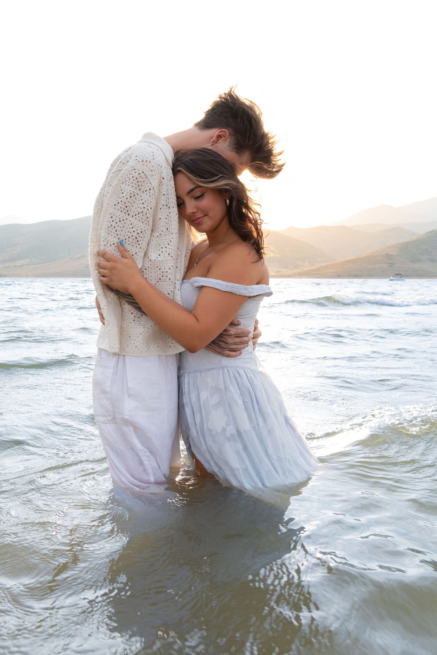 A romantic couple embracing in the ocean waves at sunset wearing white and blue outfits at the beach.
