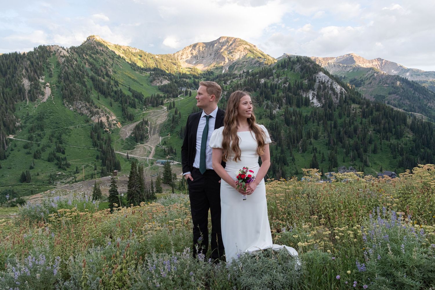 Bride and groom pose together in a scenic mountain landscape with blooming wildflowers and rugged peaks.