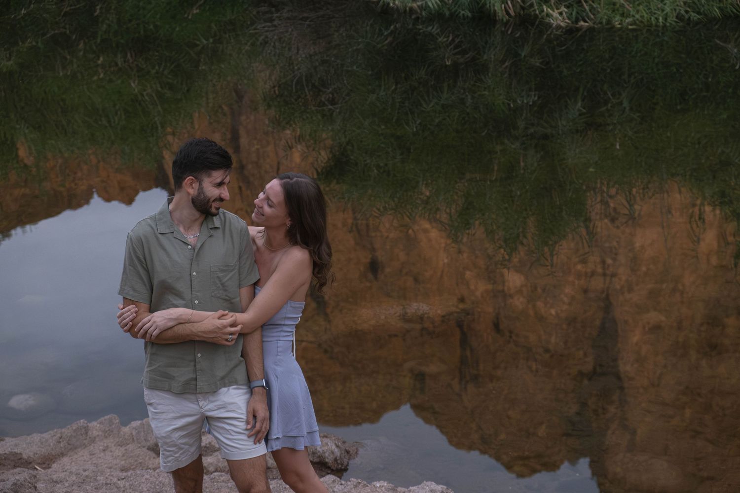 A couple shares a romantic moment by a lake with mountain reflections at sunset.
