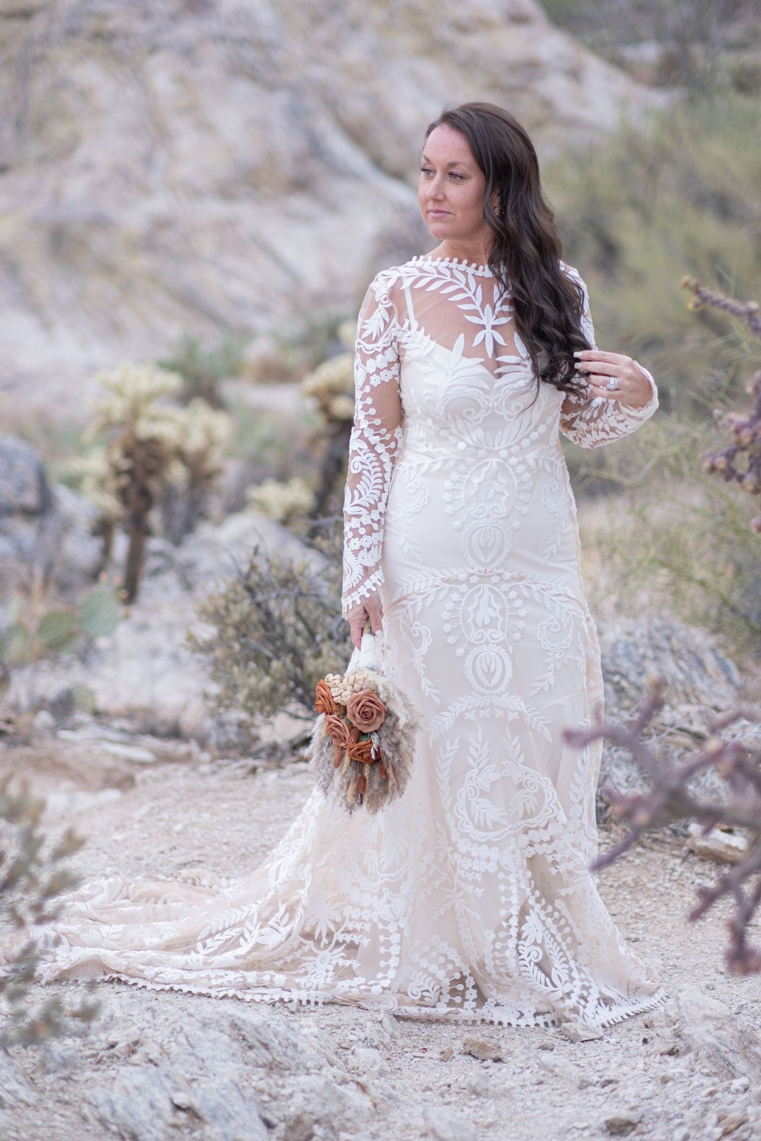 A bridal elopement portrait in a desert setting features an elegant lace wedding dress with sheer long sleeves and flowing train.