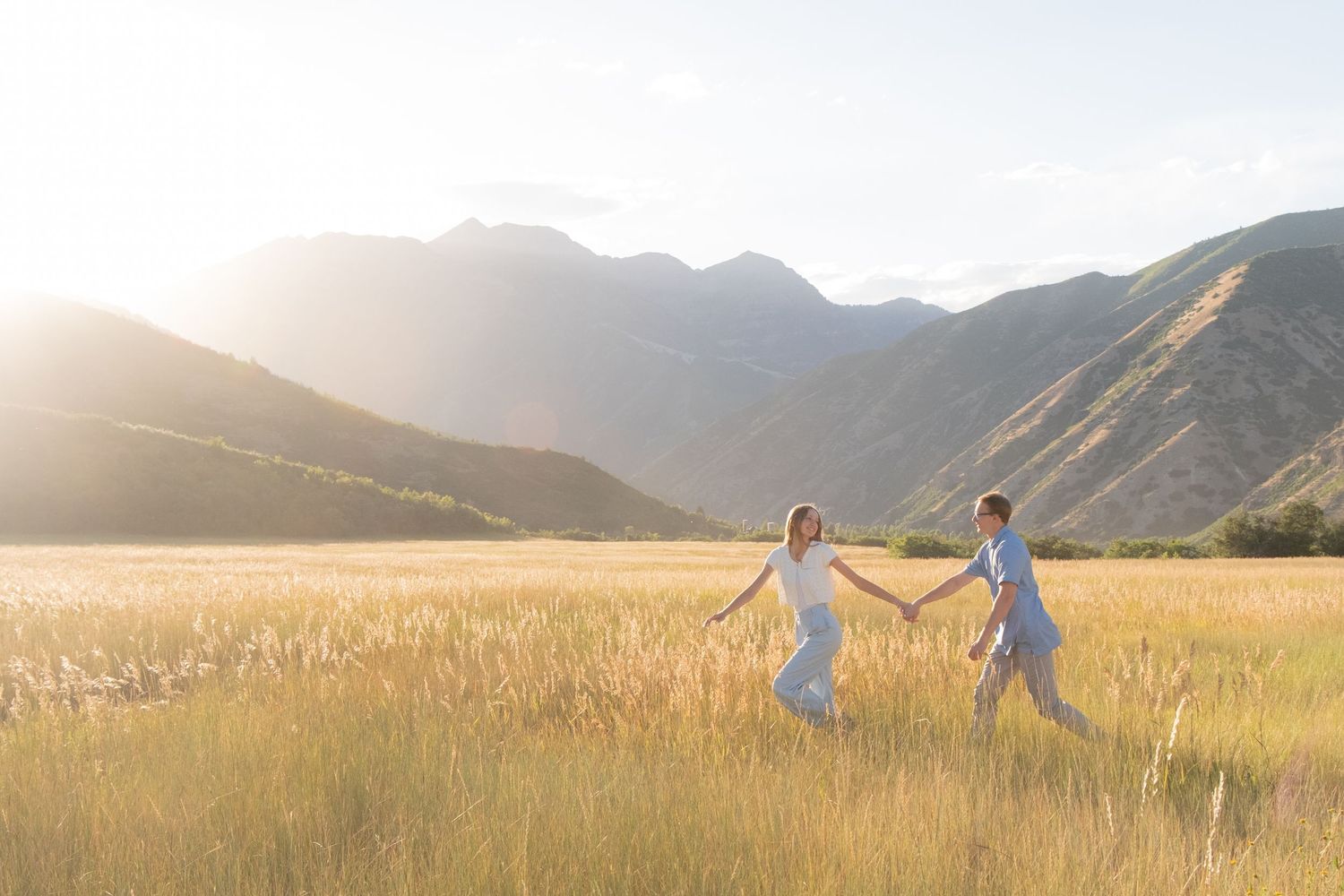 Young couple runs through golden wheat field at sunset with mountains in background.