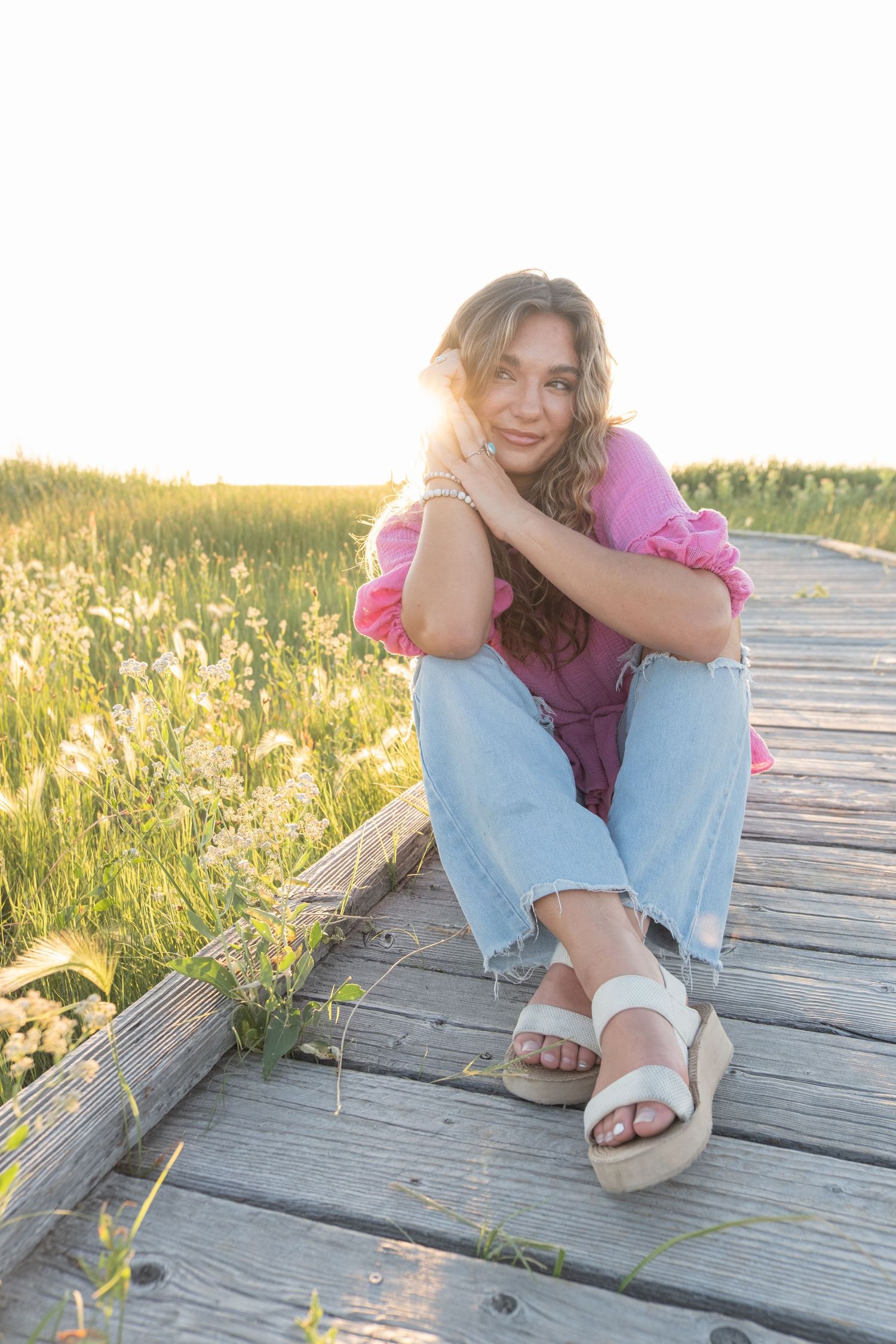 Someone wearing light blue jeans and sandals sits on a wooden boardwalk surrounded by yellow wildflowers at sunset.