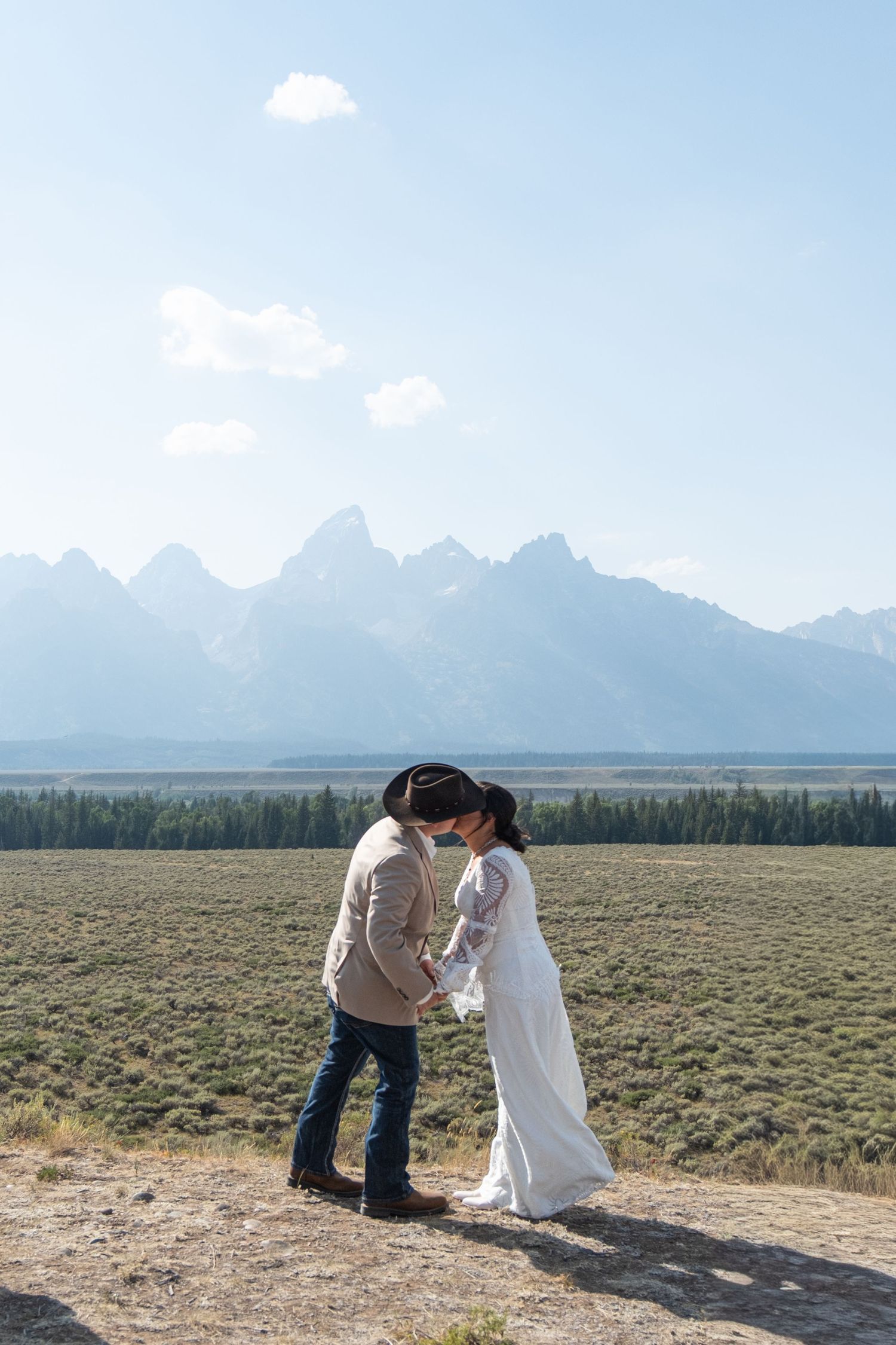Couple sharing a kiss in wedding attire with cowboy hats, Grand Teton mountains backdrop, open field, sunny day.