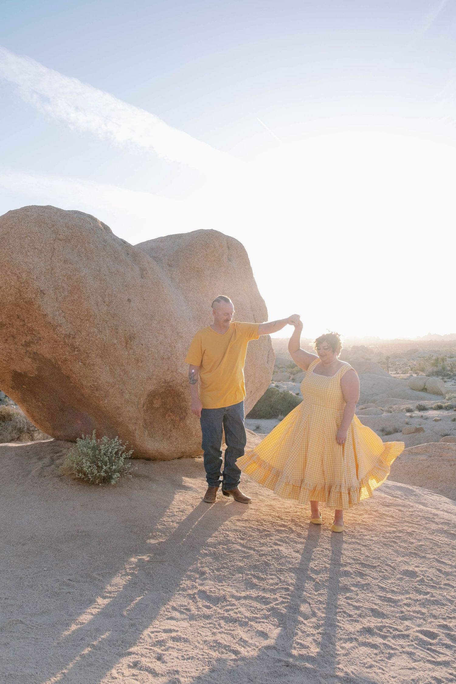 A couple in matching yellow outfits dances together among desert boulders at sunset.