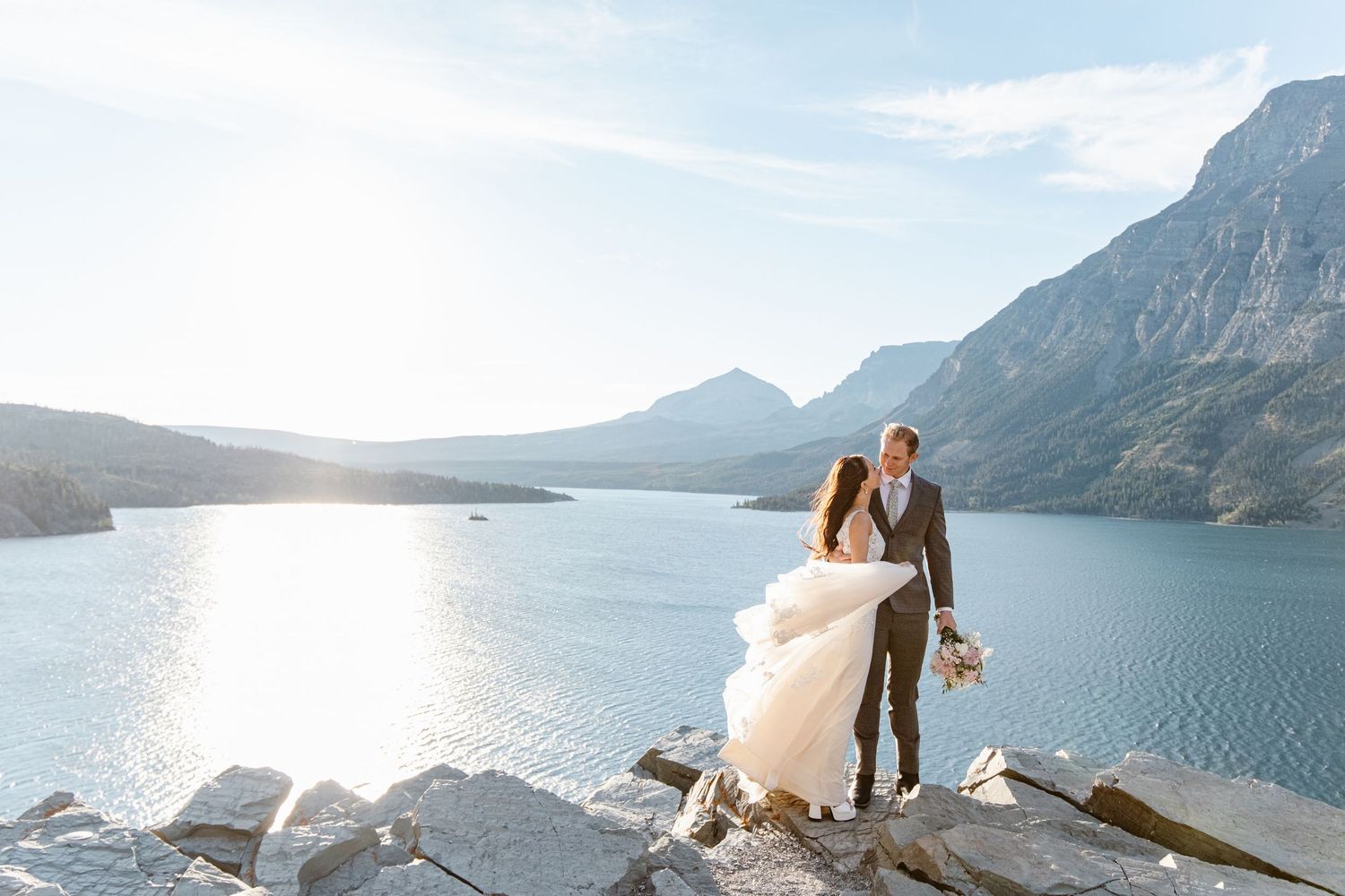 A couple shares a romantic moment on a mountain cliff overlooking a stunning lake and mountain landscape at sunset.