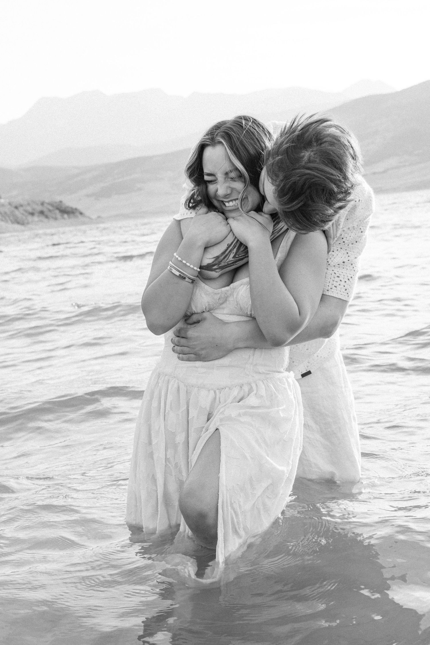 Black and white photo of a couple embracing while standing in ocean waves wearing white clothing at sunset.