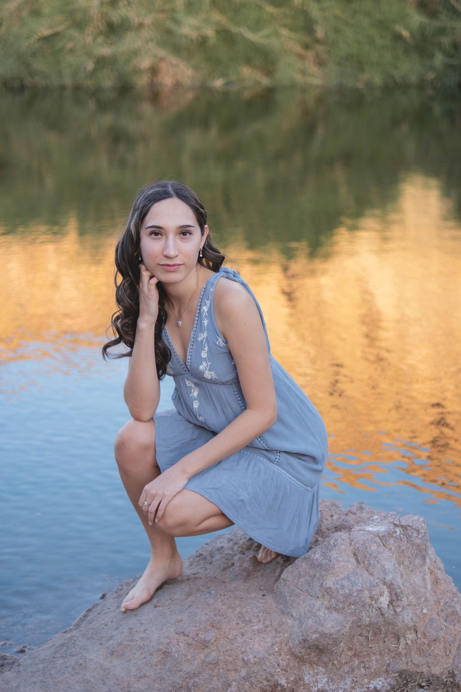 A person poses on a rocky outcrop beside a calm lake at sunset.