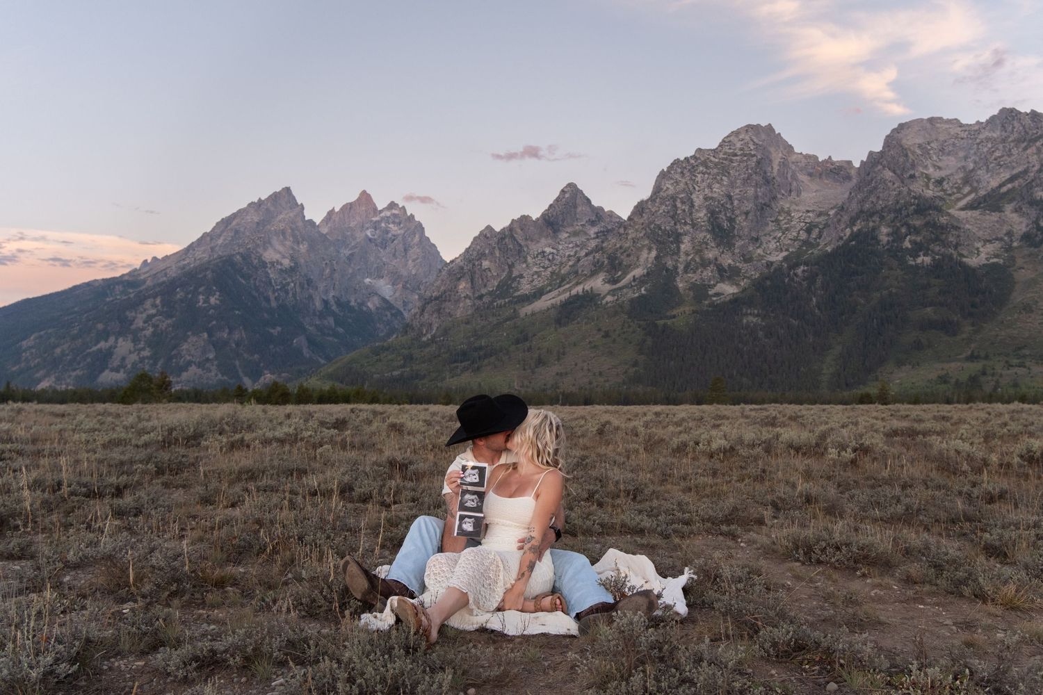 Couple sitting together in a grassy field with the Grand Teton mountain range at sunset.
