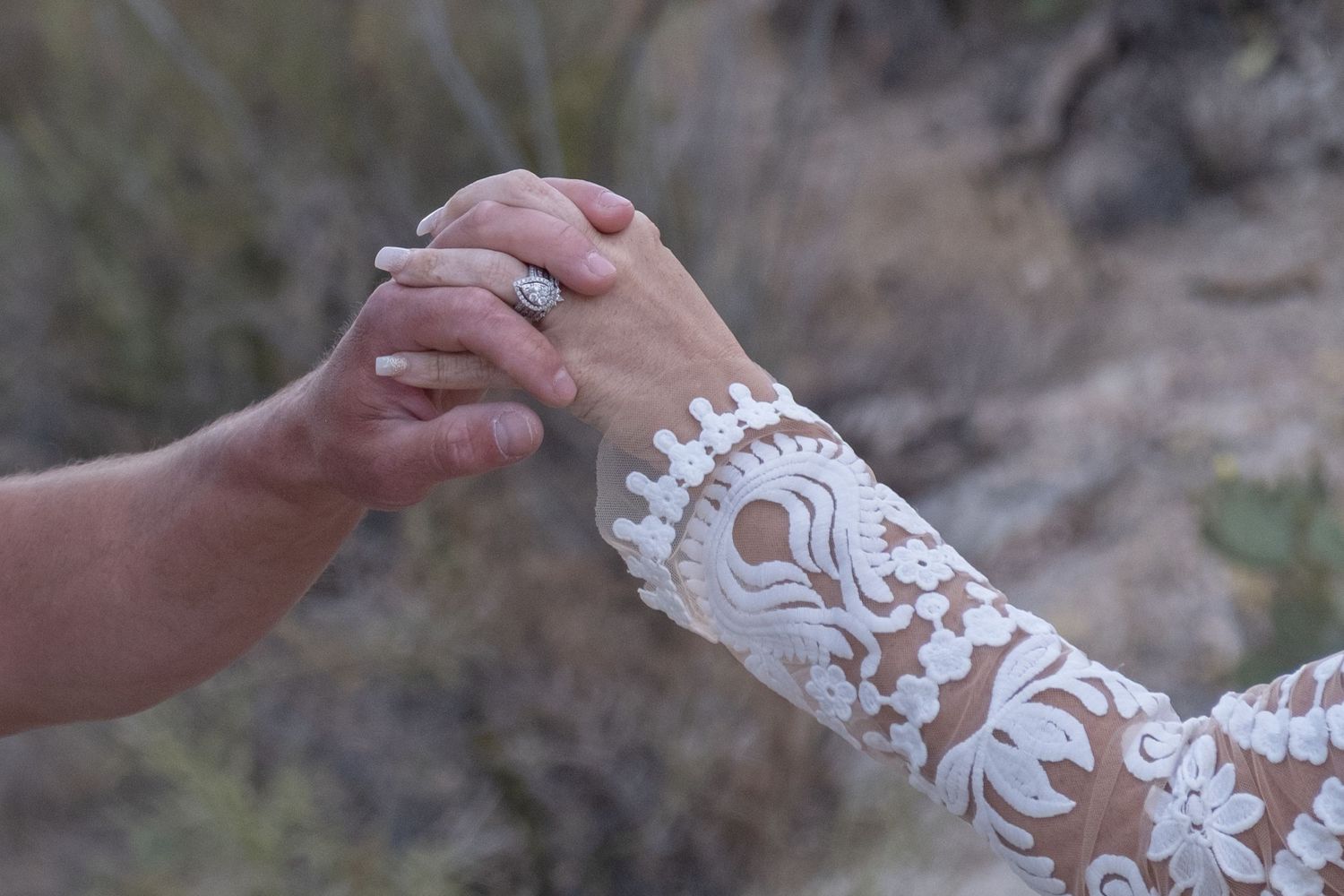 A close-up photo showing clasped hands with a white lace bridal sleeve detail against a blurred outdoor background during their elopement.