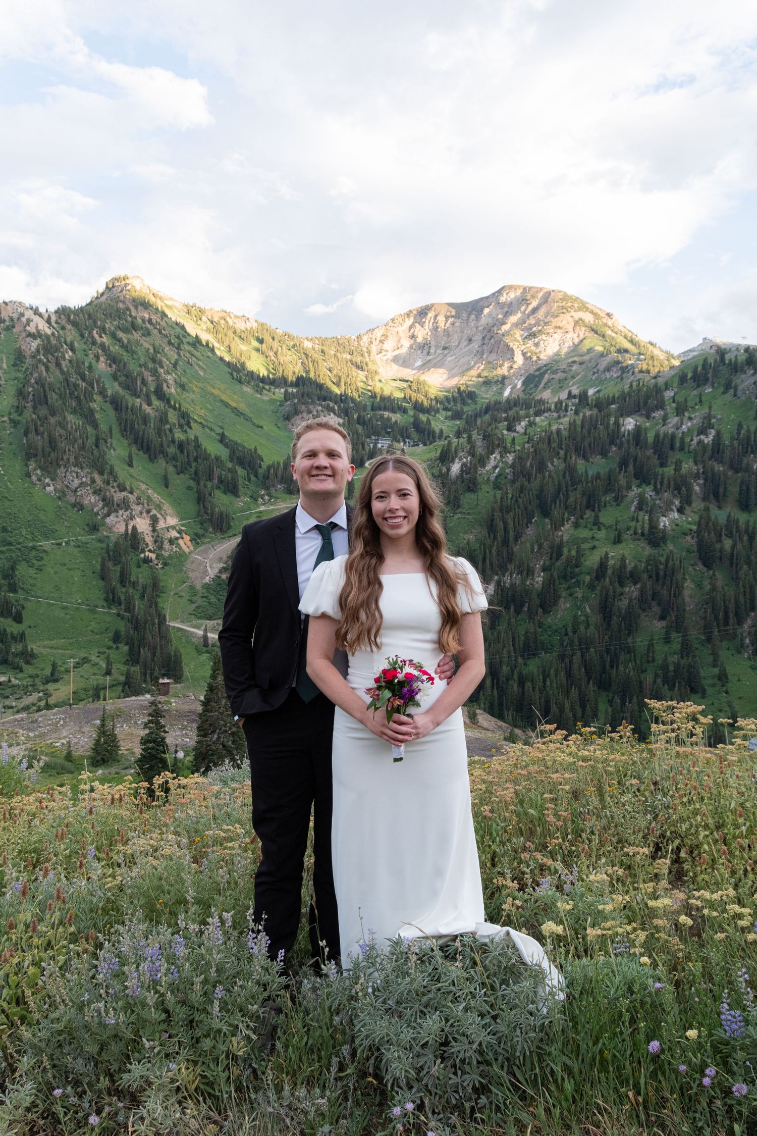 Wedding couple poses in flower-filled mountain meadow with majestic snow-capped peaks in background.