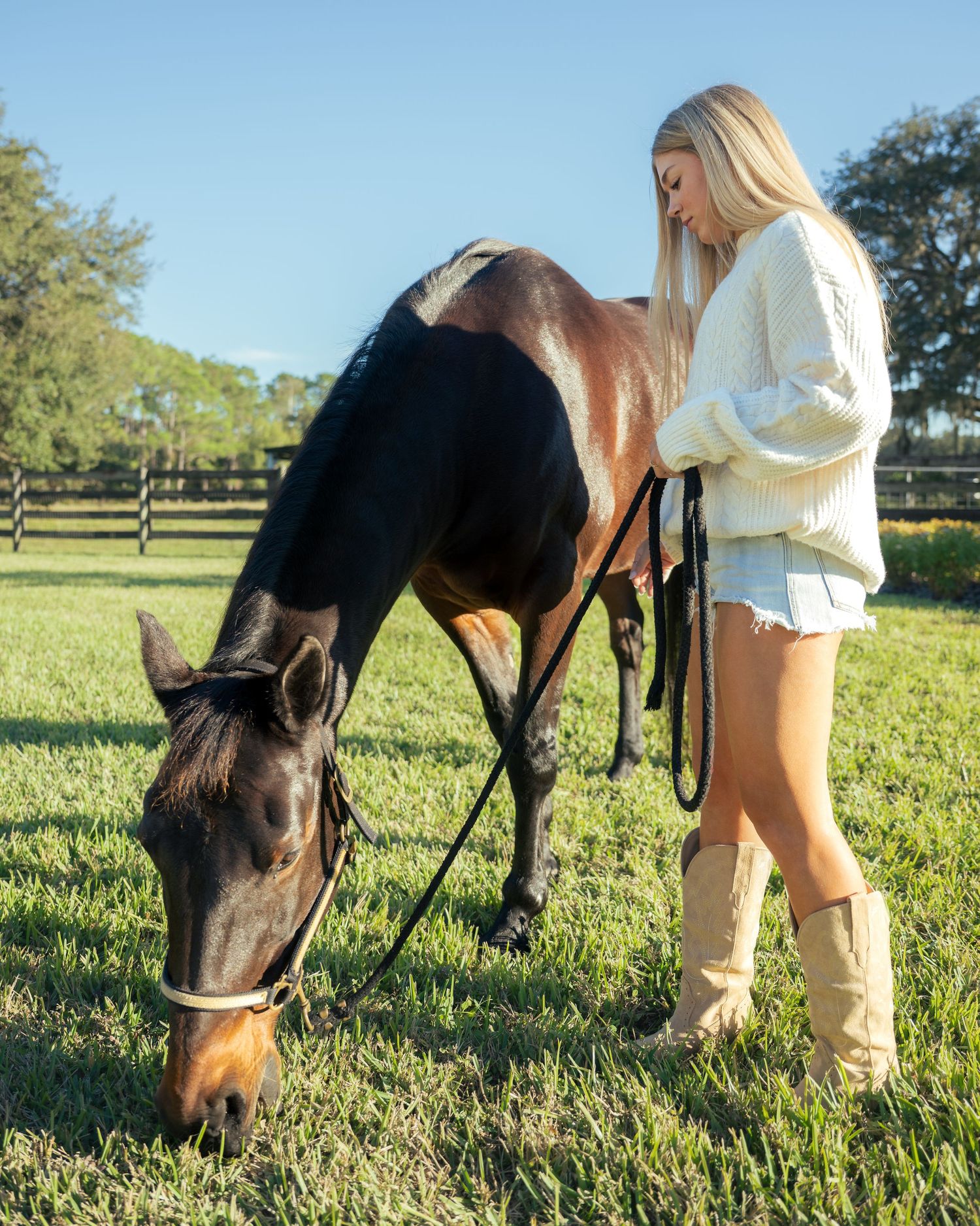 Blonde woman in cream sweater and cowboy boots standing with a bay horse on green pasture.