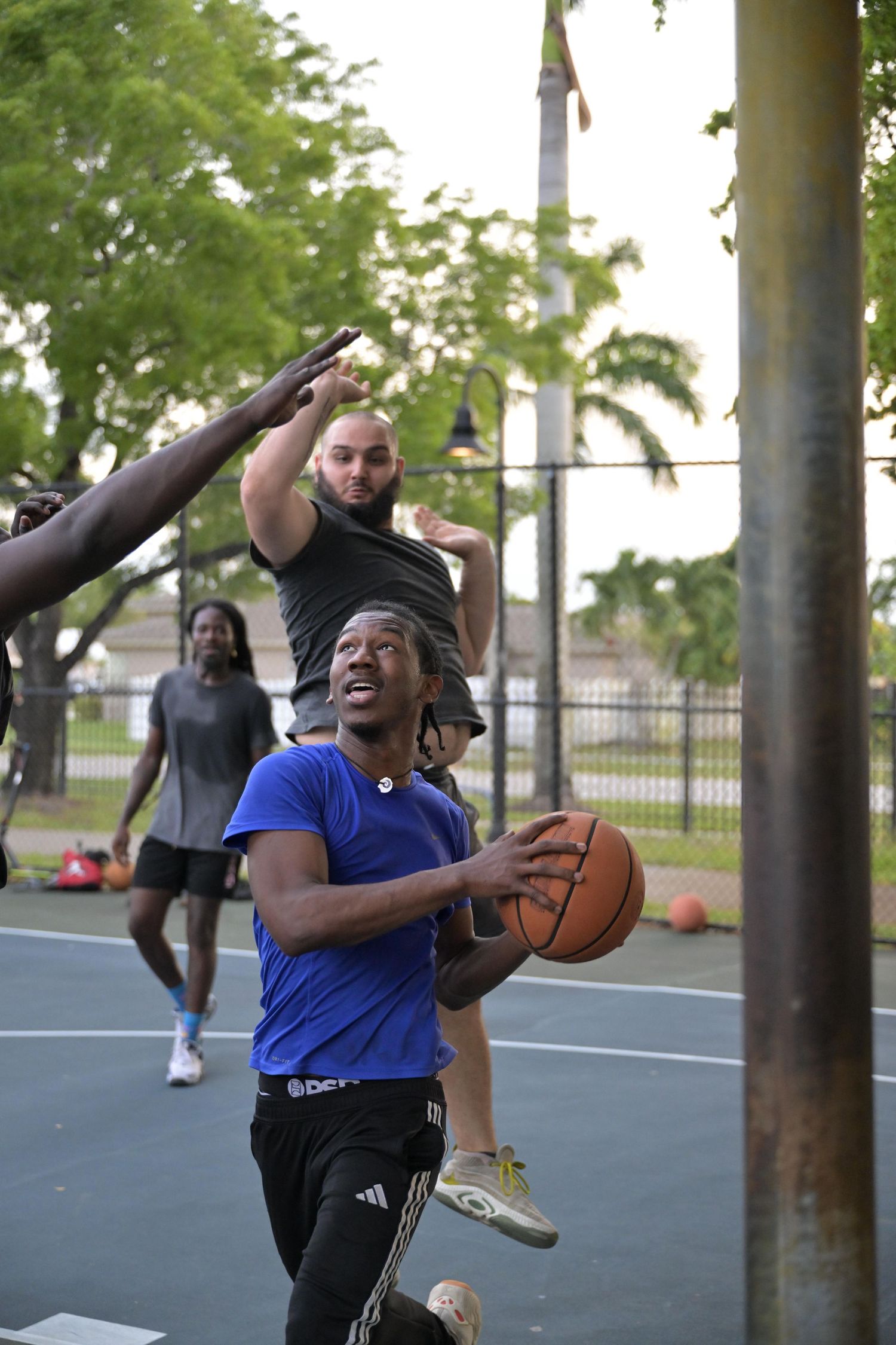 Players compete in an outdoor basketball game, with one player driving to the basket while defenders attempt to block the shot.