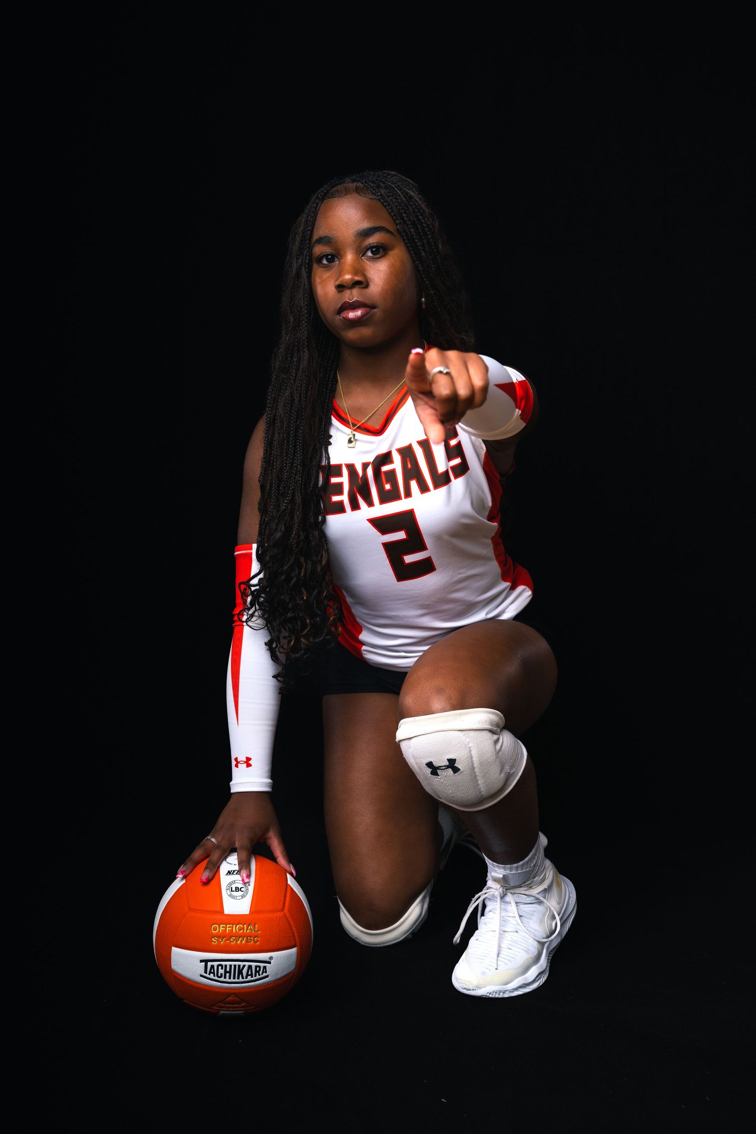 Female volleyball player in Bengals number 2 jersey kneels with Tachikara ball, pointing at camera against black background.