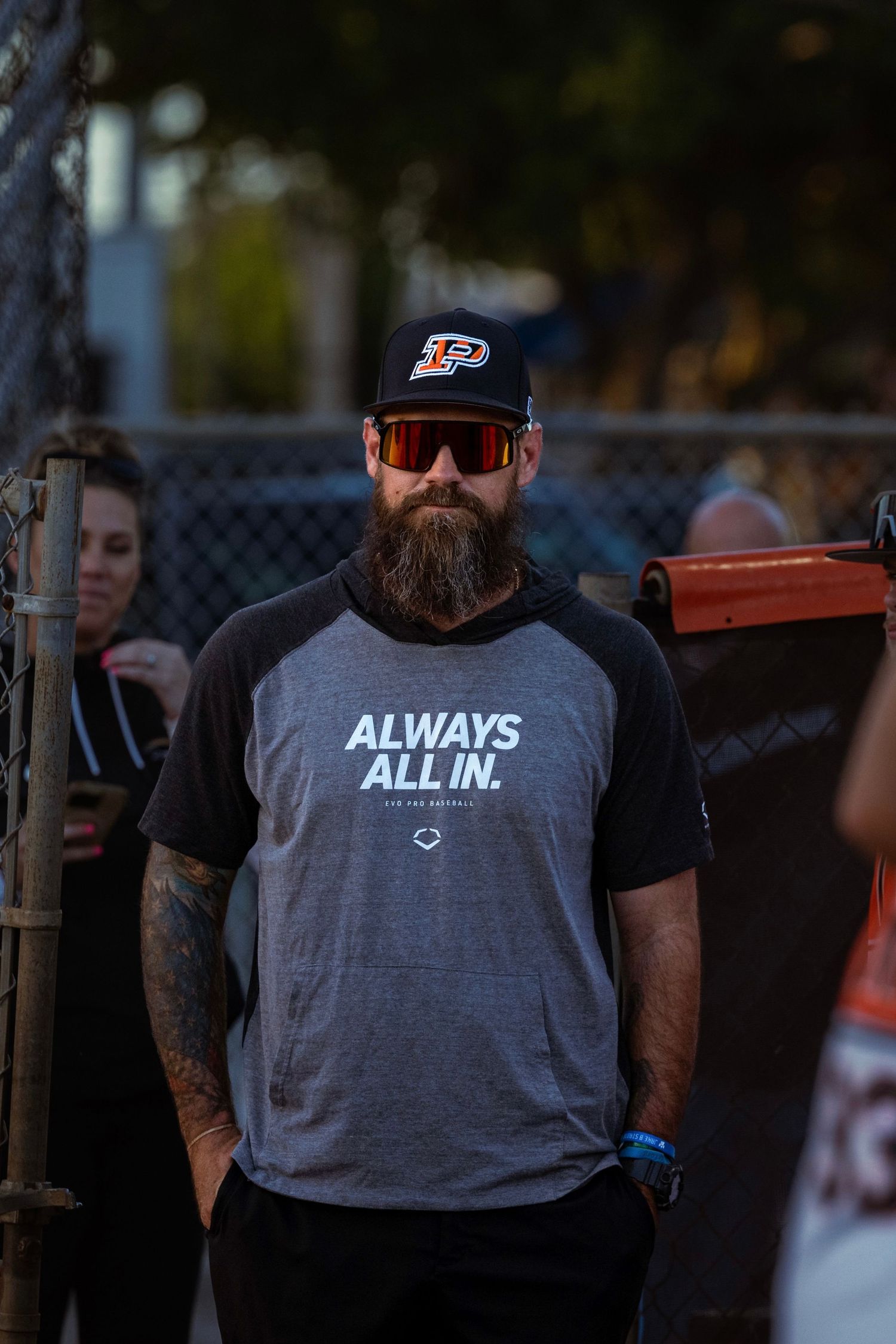 Portrait of a coach wearing a 'Always All In' shirt and cap, leaning on a fence in Fort Lauderdale, reflecting commitment.