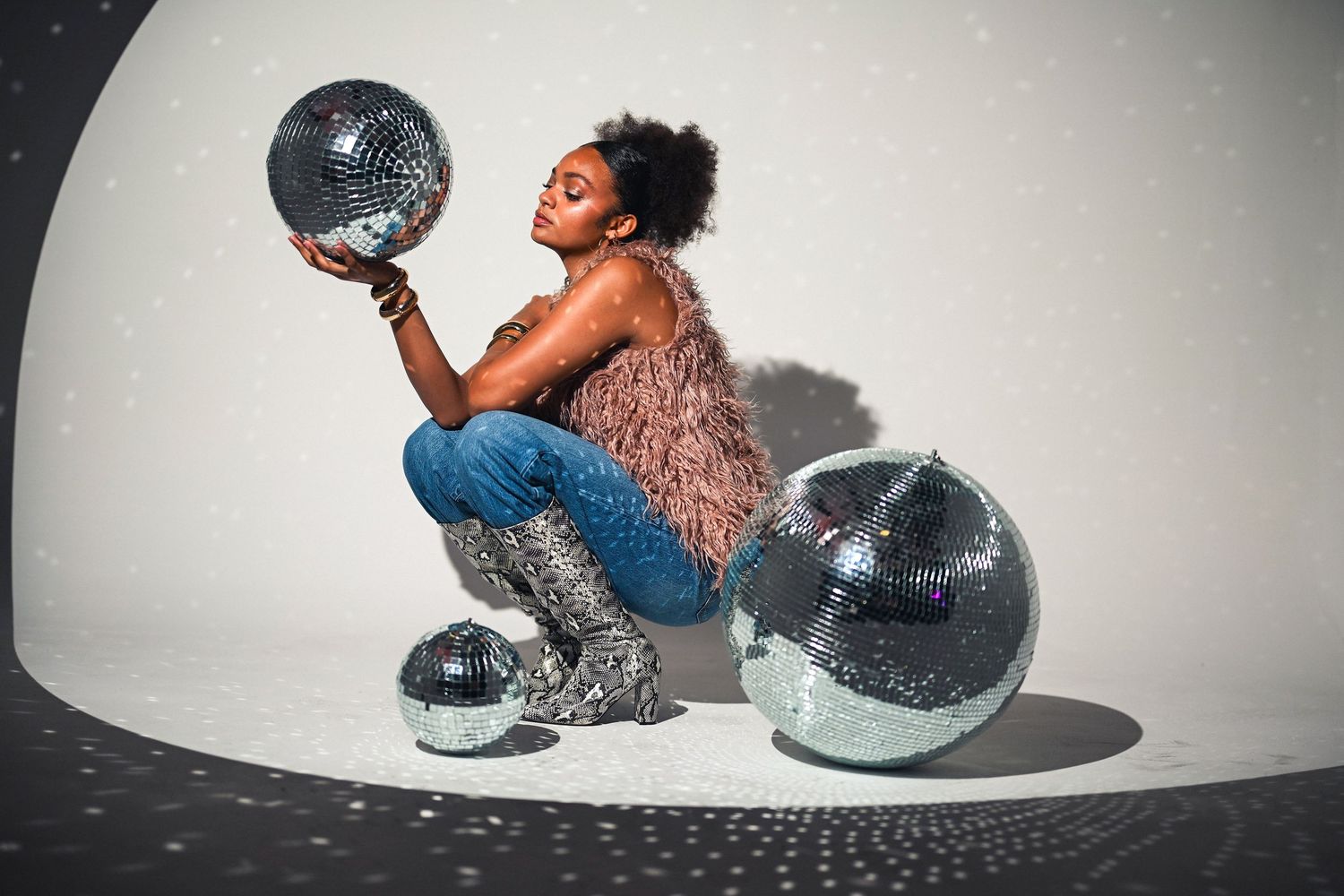 A stylish woman crouches among large mirrored disco balls in a studio setting wearing jeans and a fur vest.