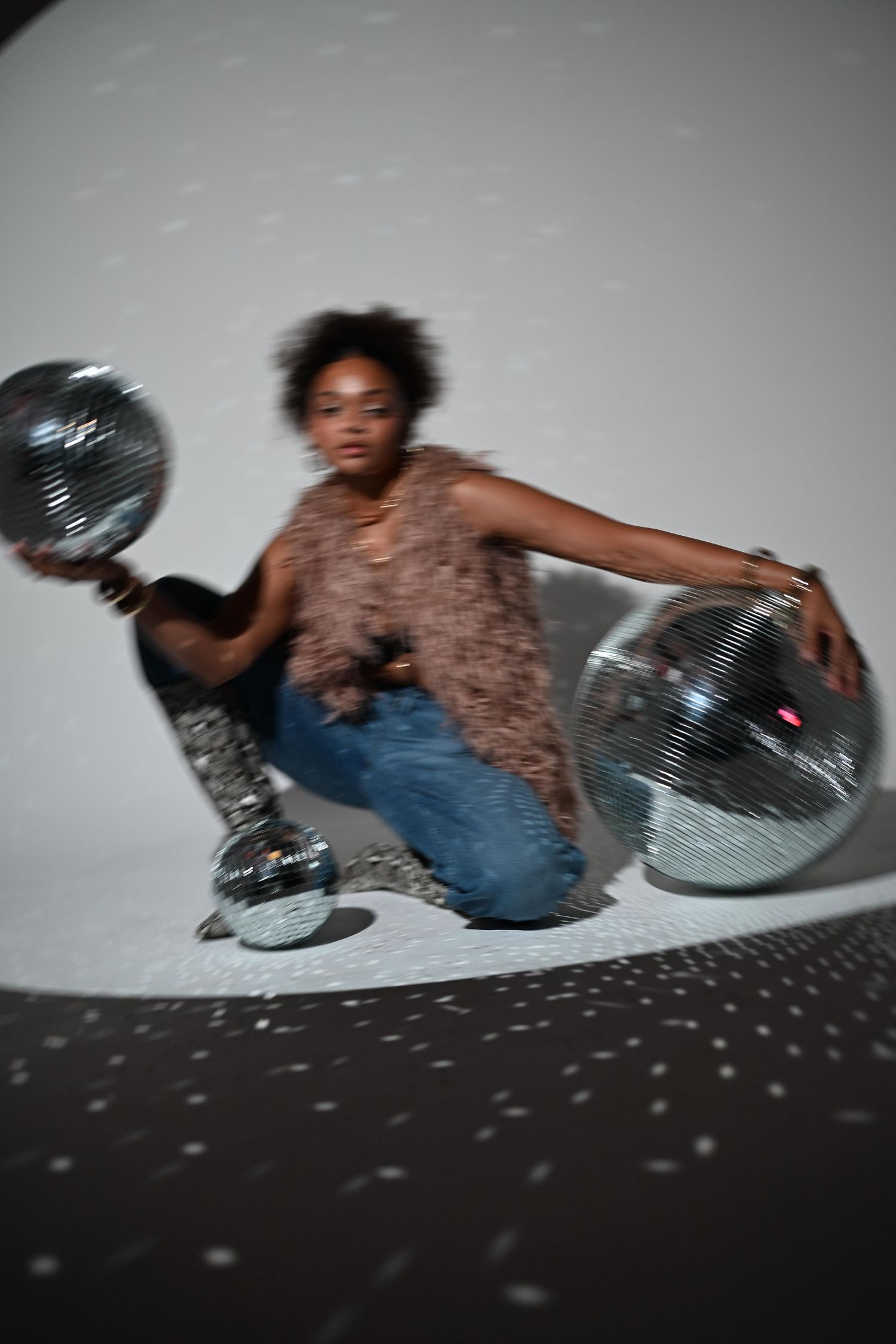 Black woman in fur vest and jeans poses dynamically with silver disco balls under studio spotlight.