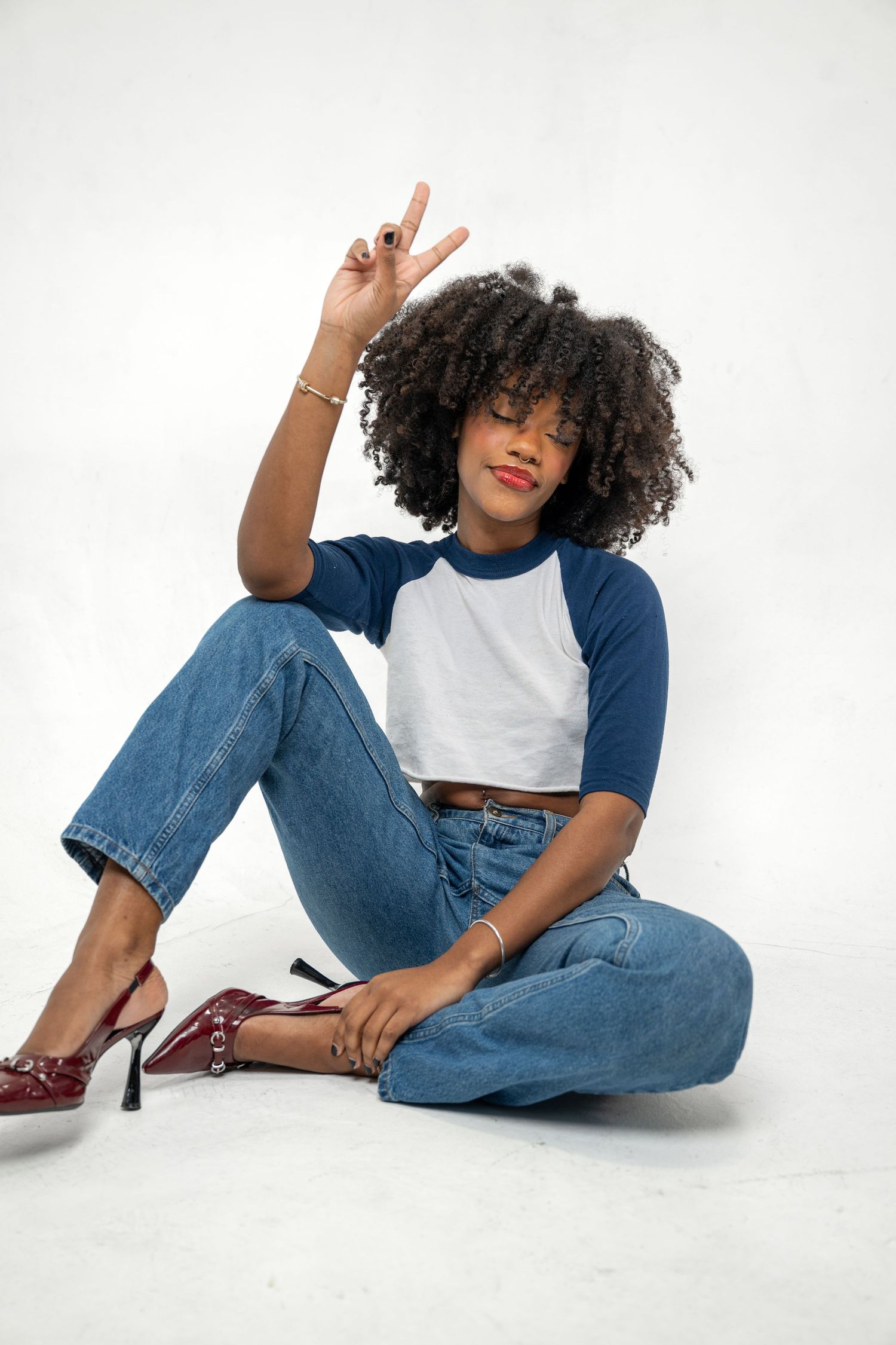 Confident young Black woman in jeans and raglan tee giving peace sign while seated on white floor.